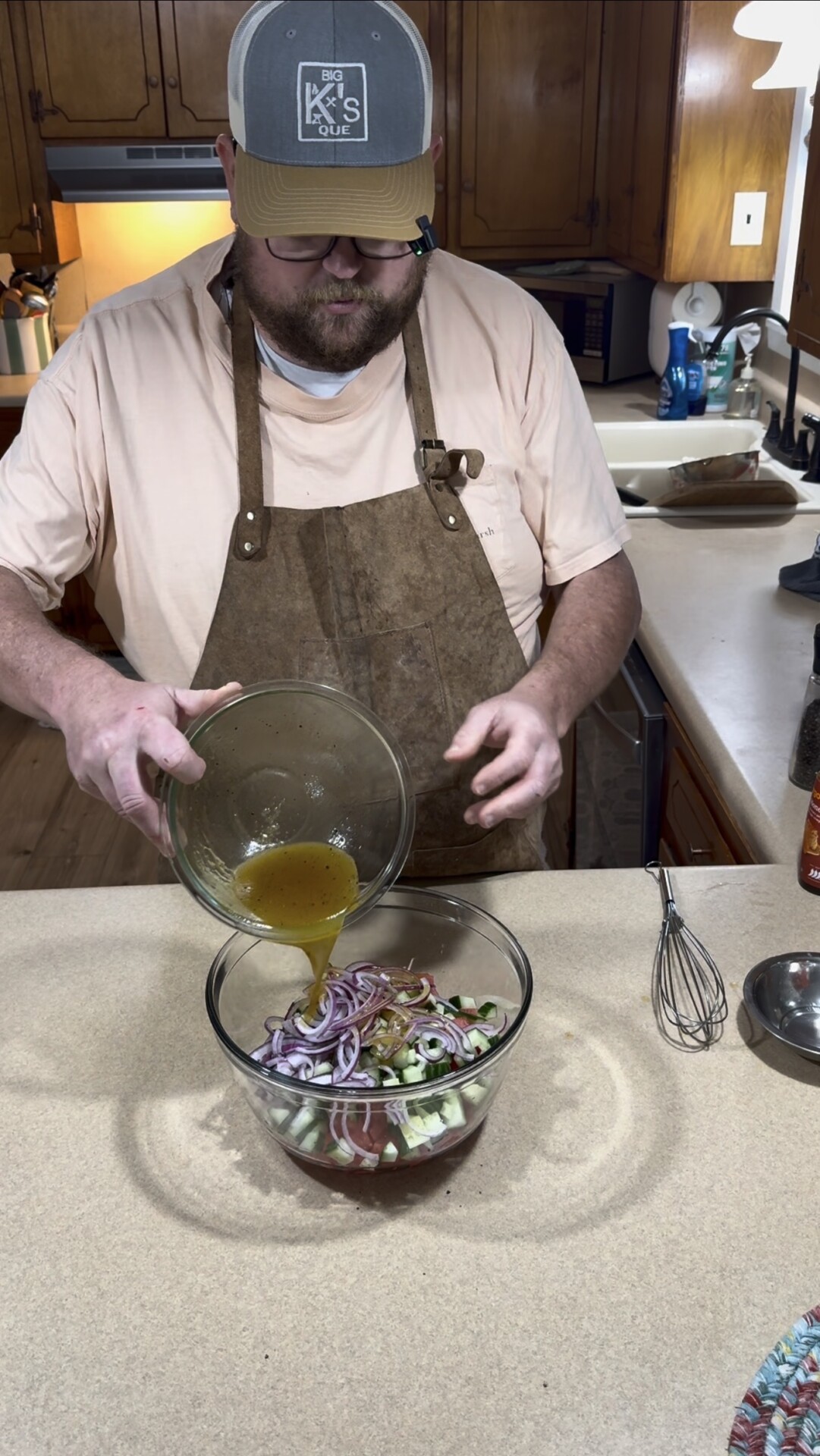 A man in an apron and cap pours dressing from a bowl onto sliced vegetables in a glass mixing bowl on a kitchen counter. A whisk and other kitchen items are nearby.