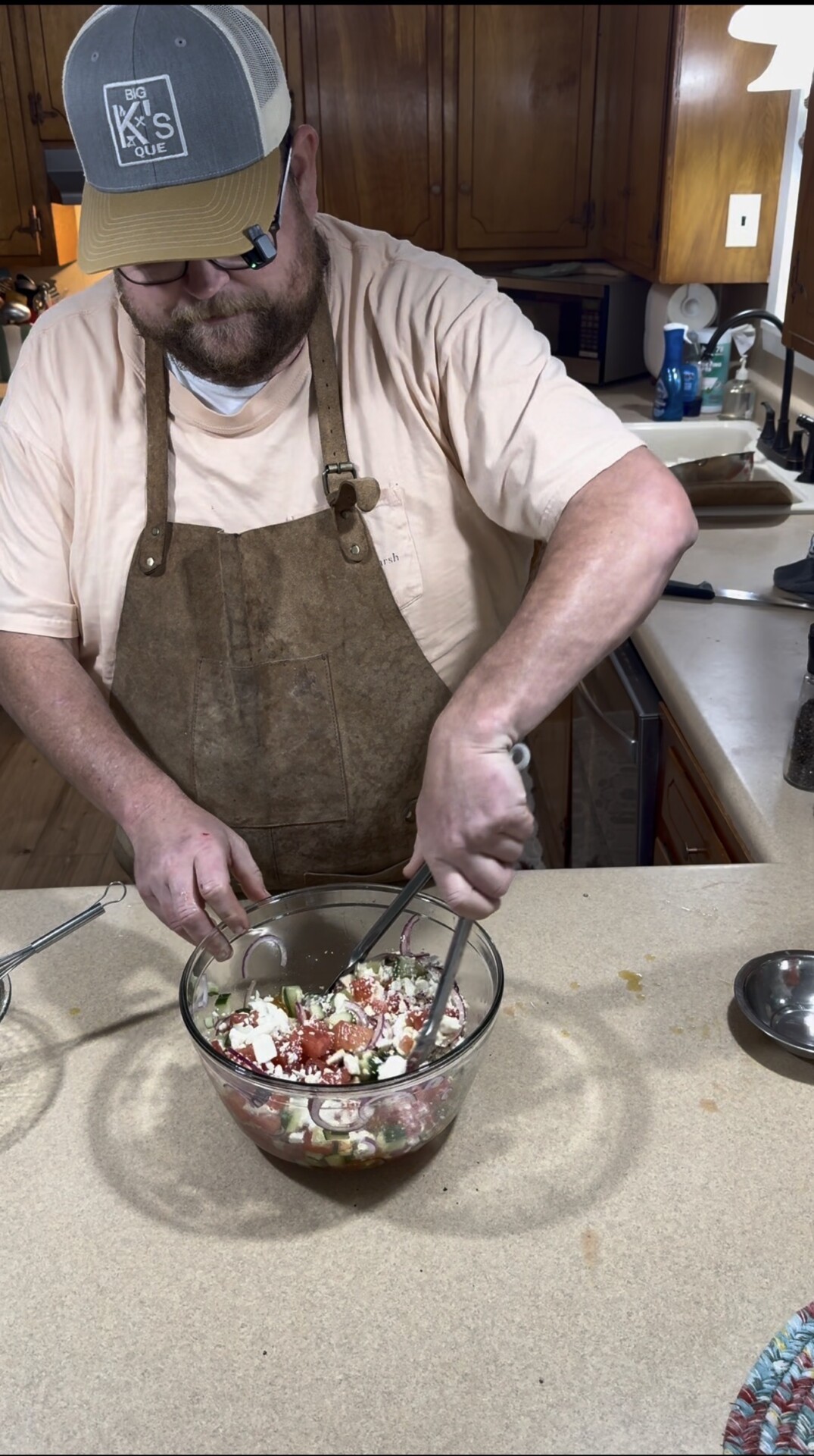 A person wearing an apron and cap is mixing ingredients in a glass bowl on a kitchen counter, using a meat chopper and fork. The kitchen has wooden cabinets and various utensils visible in the background.