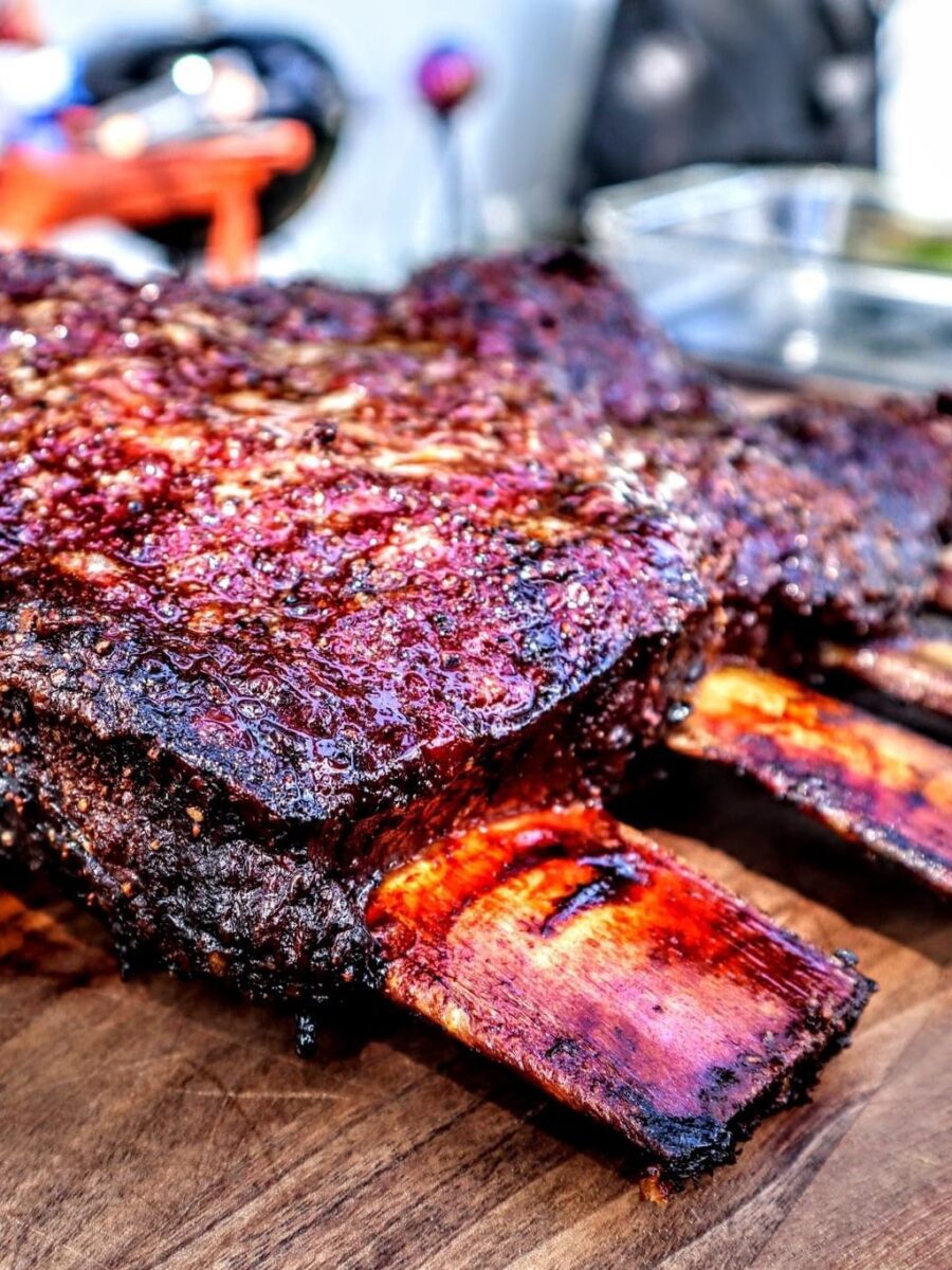 Close-up of juicy, charred beef ribs resting on a wooden cutting board, showing a crispy, caramelized crust and exposed bones.