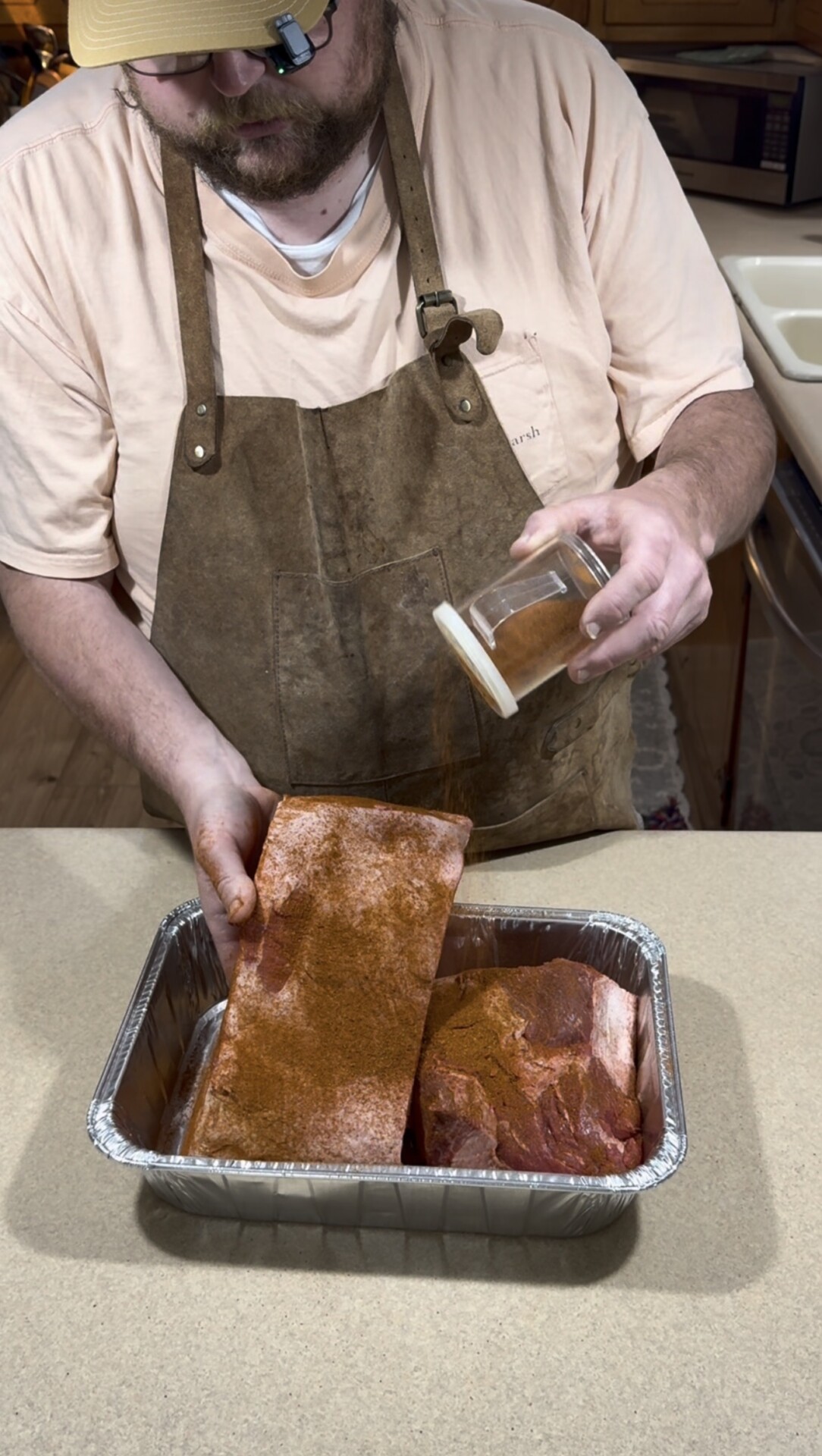 A person wearing a brown apron sprinkles seasoning from a jar onto slabs of meat in a foil tray on a kitchen counter.