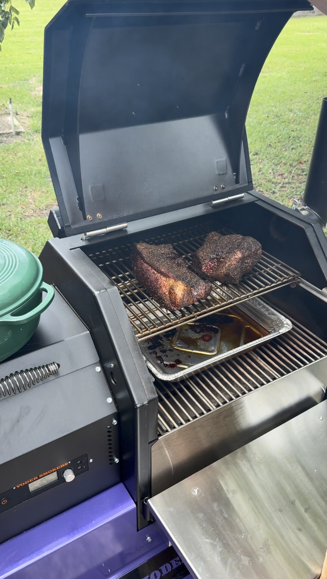 Two large pieces of seasoned meat are smoking on a grill rack above a drip pan, with a green pot nearby and a grassy yard in the background. The grill lid is open, showing the meat and equipment.