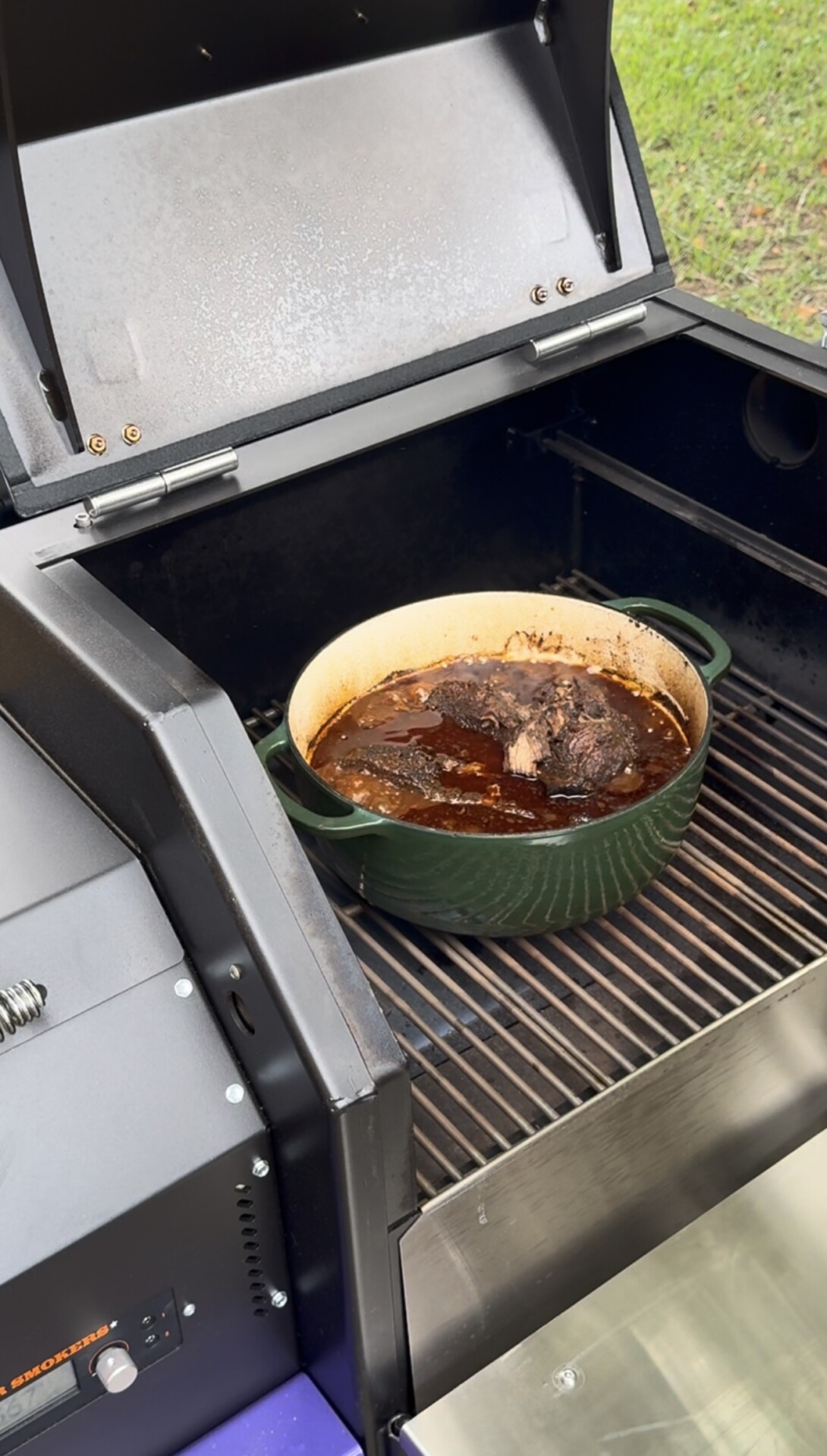 A green Dutch oven filled with braised meat and sauce sits on the grate inside an open outdoor grill, with grass visible in the background.