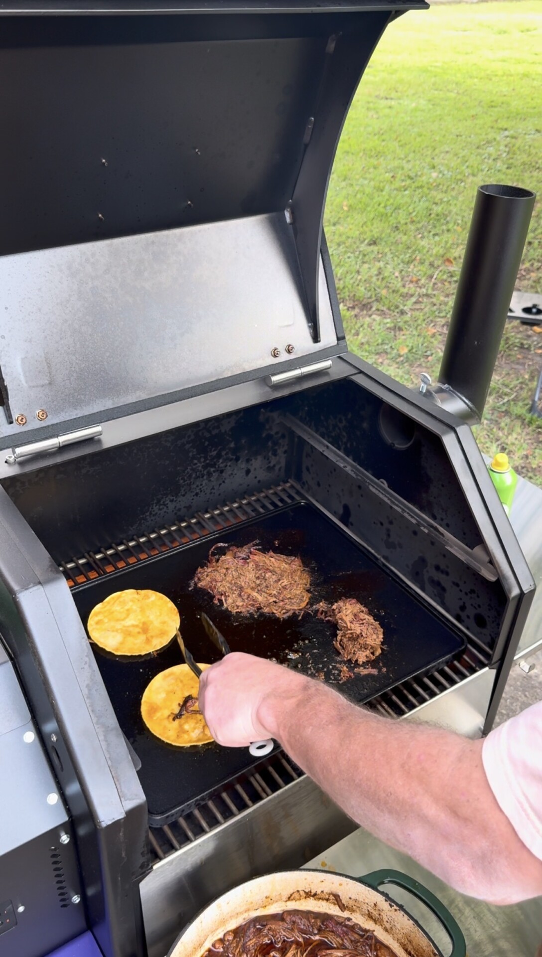 A person grills shredded meat and two corn tortillas on an outdoor barbecue grill, with a grassy lawn visible in the background.