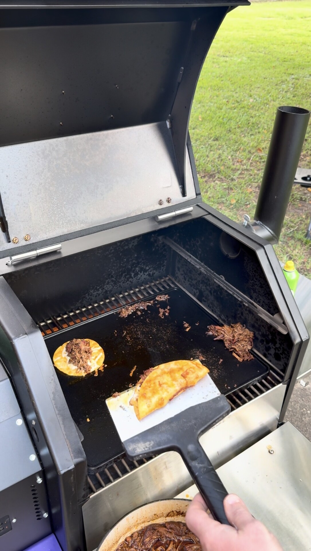 A hand holds a spatula lifting a taco from a grill. Another taco and some shredded meat are also on the grill. Green grass is visible in the background.