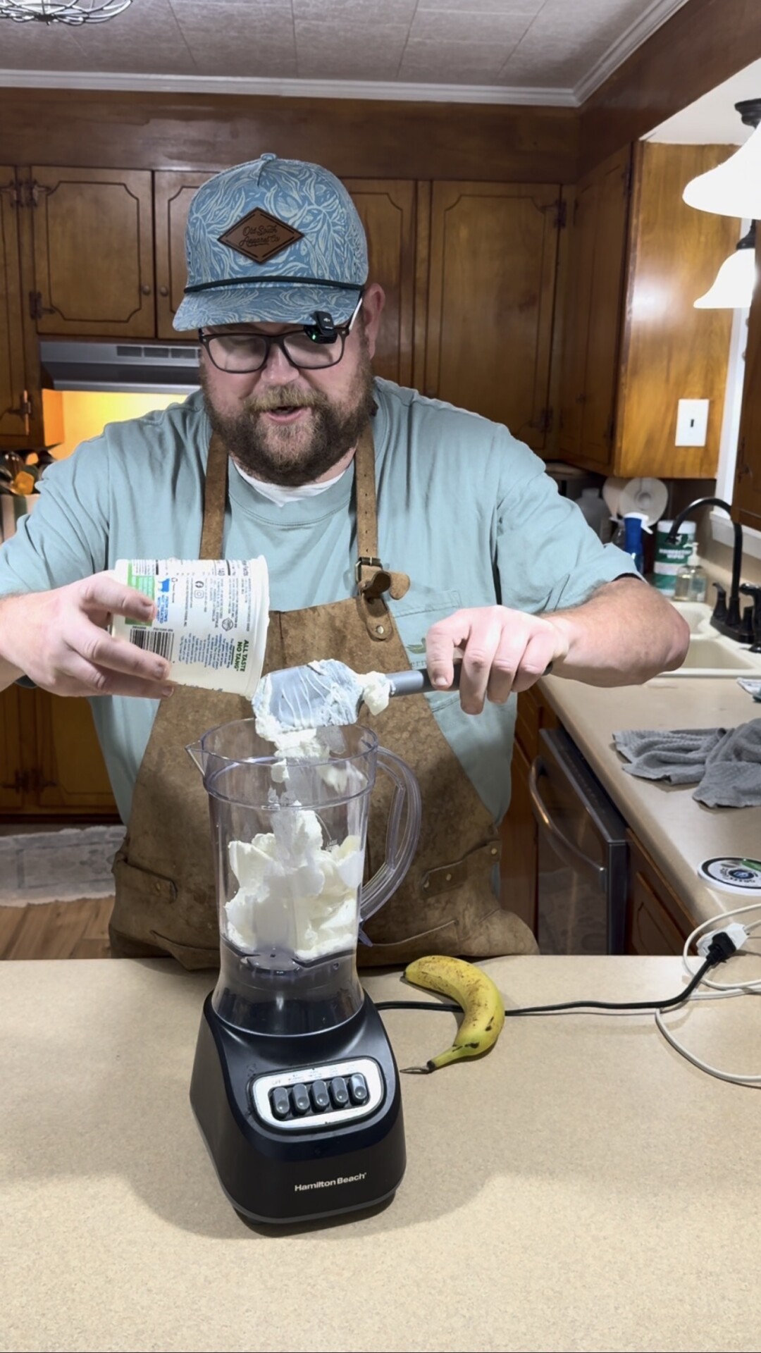 A man in an apron and hat adds a large scoop of yogurt into a blender on a kitchen counter with a banana nearby.