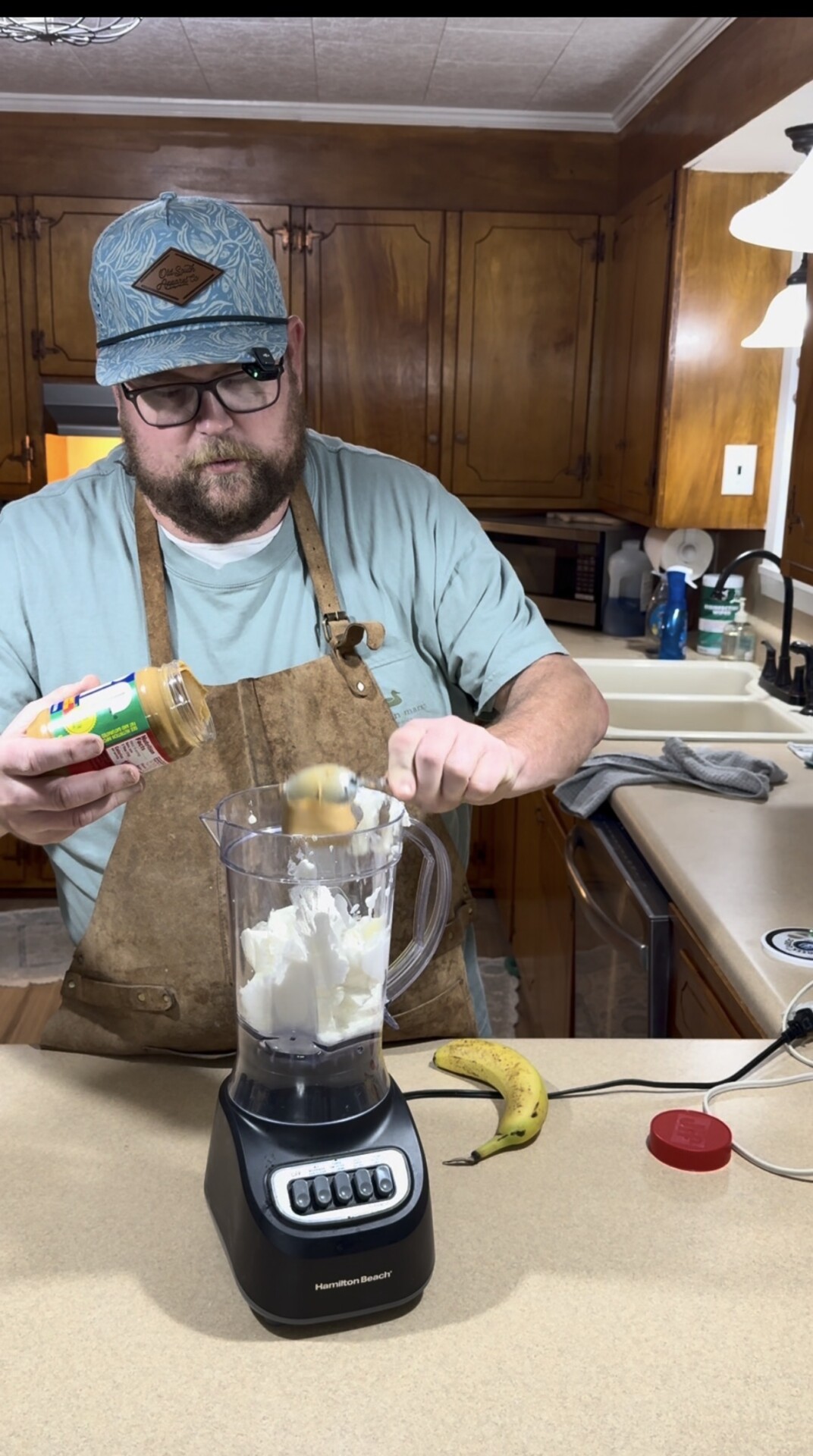 A man wearing glasses, a hat, and an apron spoons peanut butter into a blender containing a white substance, with a banana and a peanut butter jar lid on the kitchen counter.