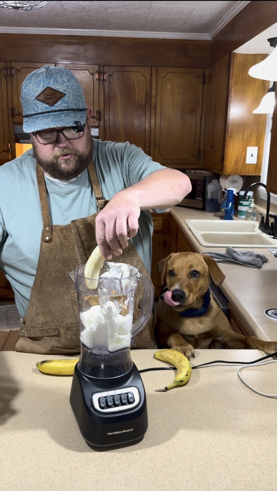 A man wearing glasses and an apron puts a banana into a blender with ice cream while a brown dog sits beside him, licking its lips. Two bananas are on the counter in a cozy kitchen.