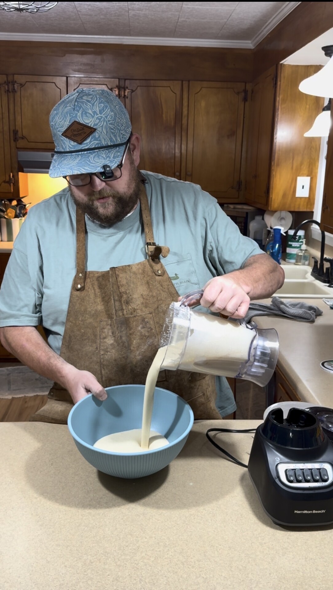 A man in a blue shirt and apron pours a blended mixture from a blender into a blue bowl in a kitchen with wooden cabinets.