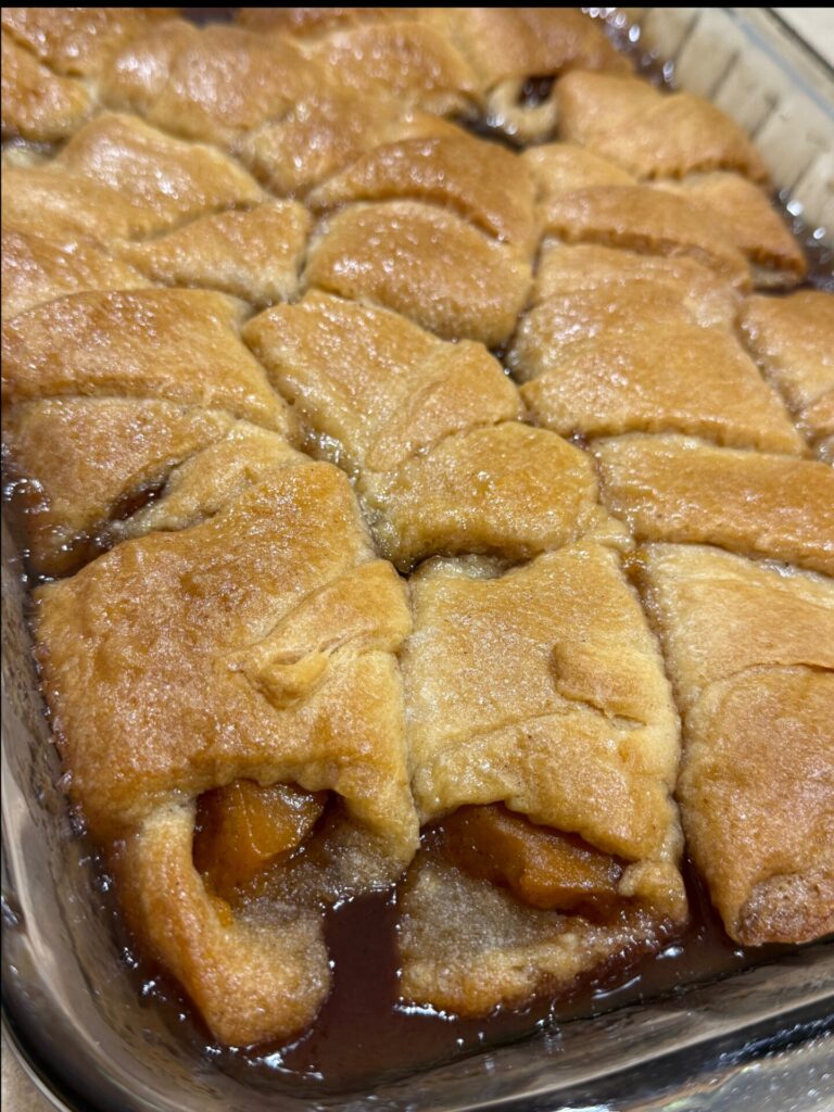 Golden-brown crescent rolls filled with baked fruit sit in a glass baking dish, surrounded by a caramelized syrup.