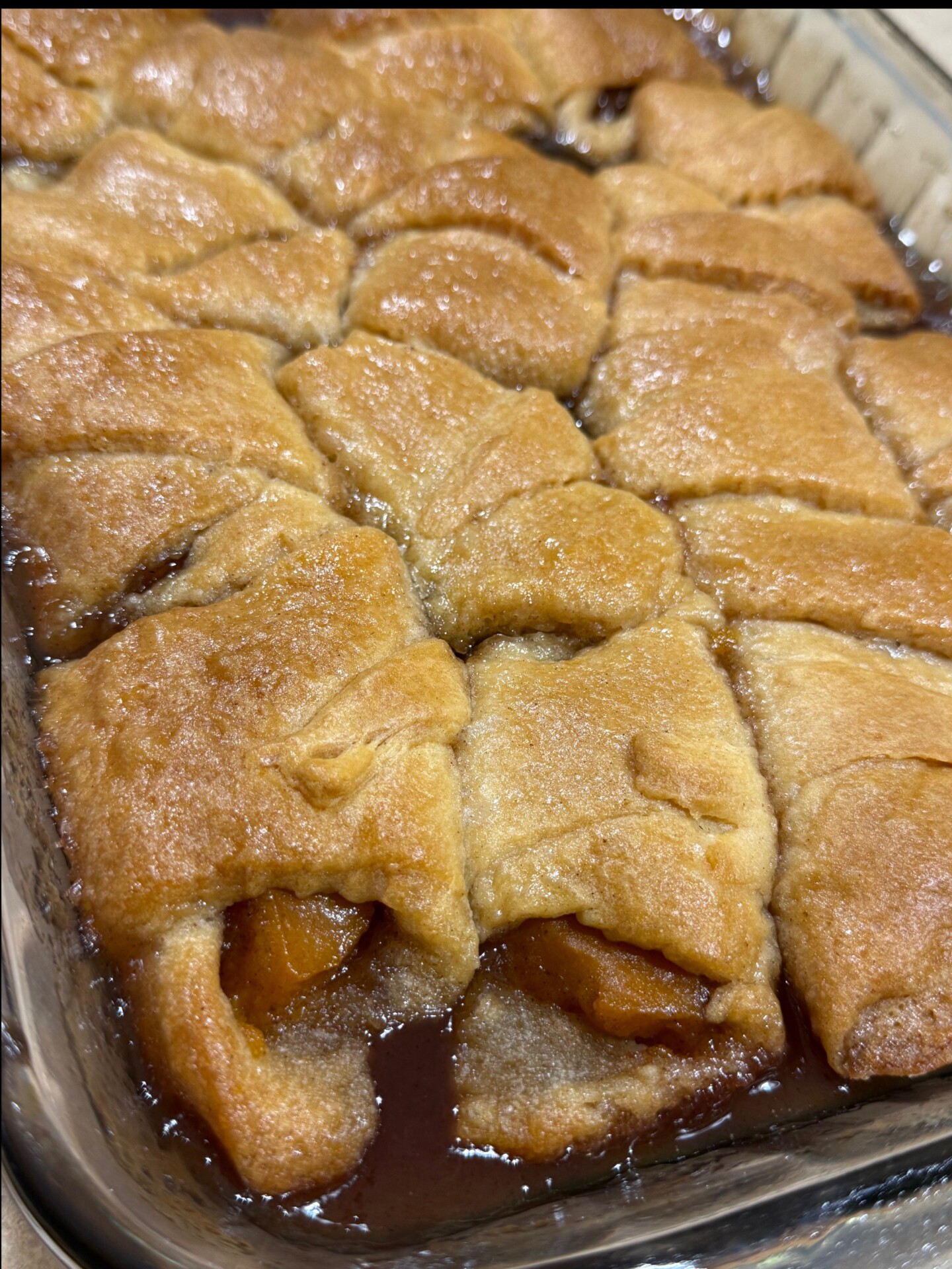 Golden-brown crescent rolls filled with baked fruit sit in a glass baking dish, surrounded by a caramelized syrup.