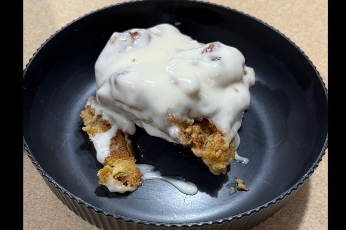 A cinnamon roll covered with creamy white icing sits in a black, round dish on a beige surface. Some crumbs and icing have dripped onto the dish.