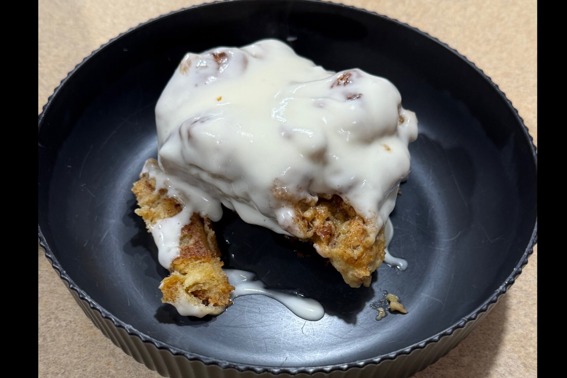 A cinnamon roll covered with creamy white icing sits in a black, round dish on a beige surface. Some crumbs and icing have dripped onto the dish.