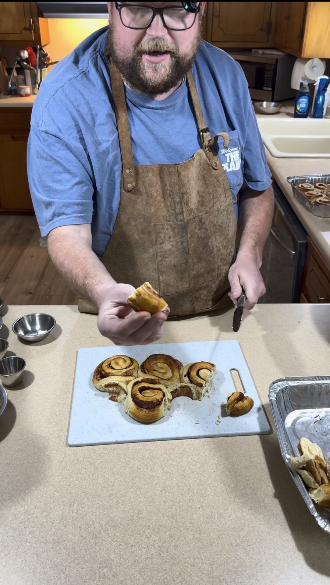 A person wearing a brown apron holds up a piece of cinnamon roll above a cutting board with more cinnamon rolls, in a kitchen setting.