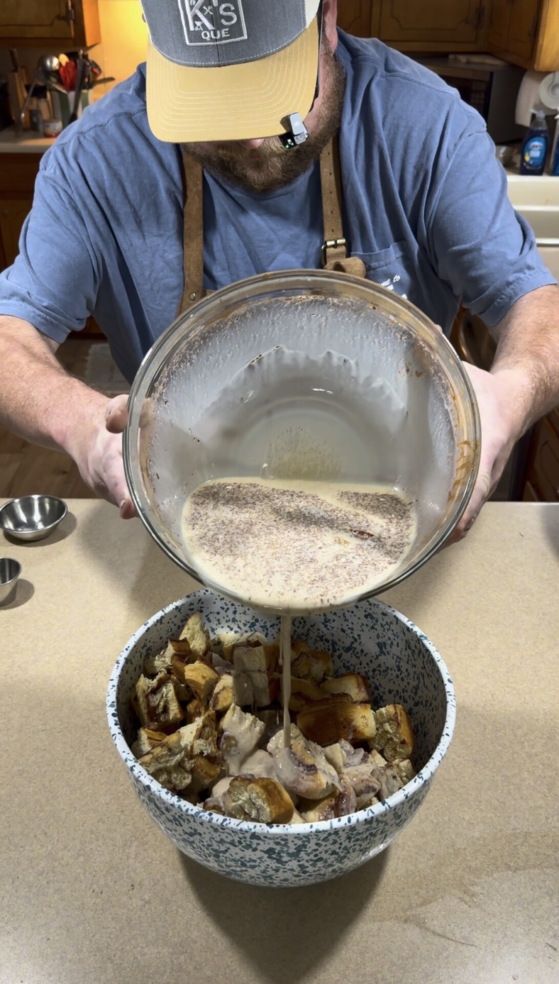 A person pours a creamy liquid mixture from a large glass bowl into a patterned bowl filled with cubed bread pieces on a kitchen counter.
