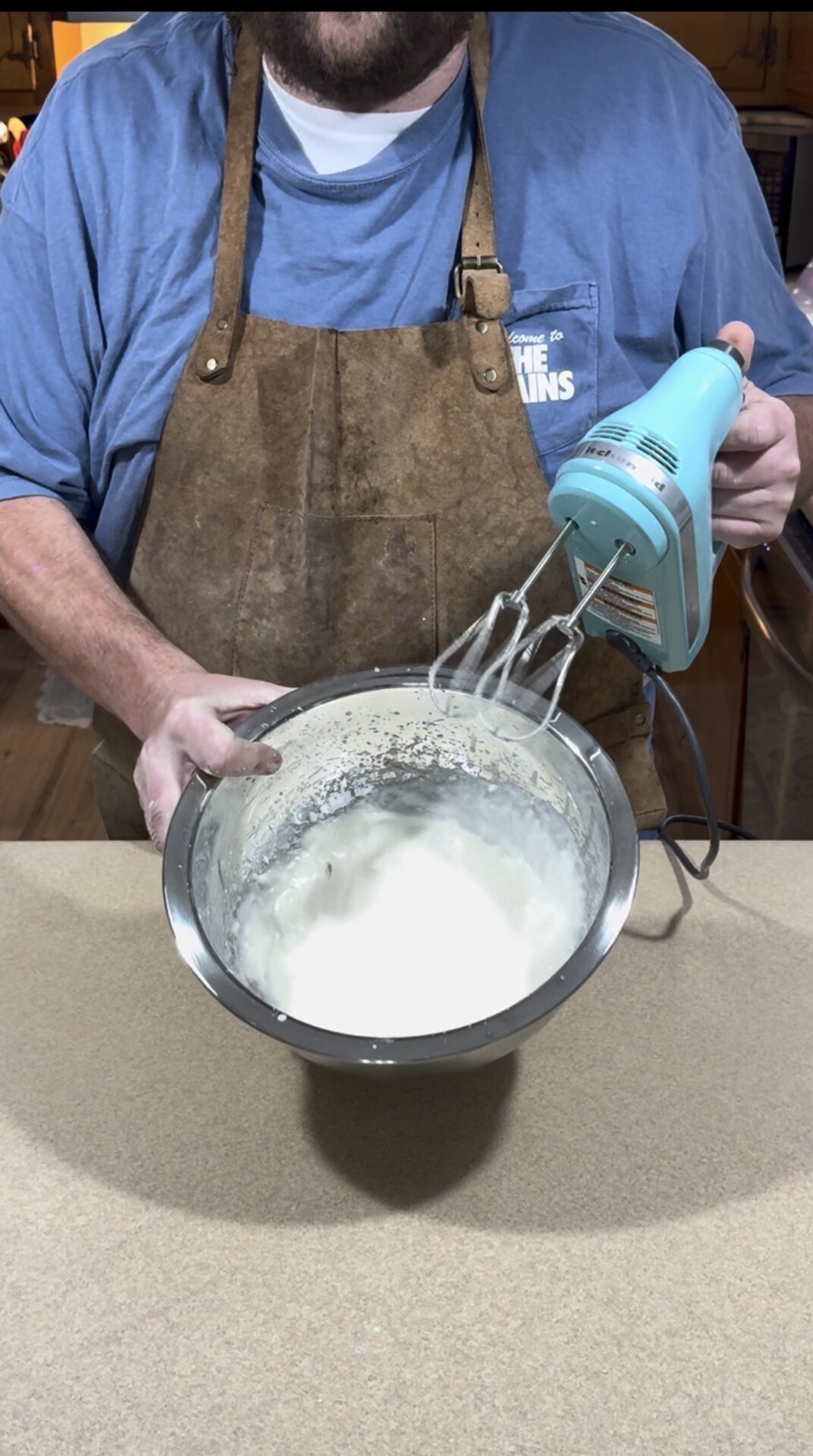 A person in a blue shirt and brown apron uses a blue electric hand mixer to whip cream in a metal bowl on a kitchen counter.