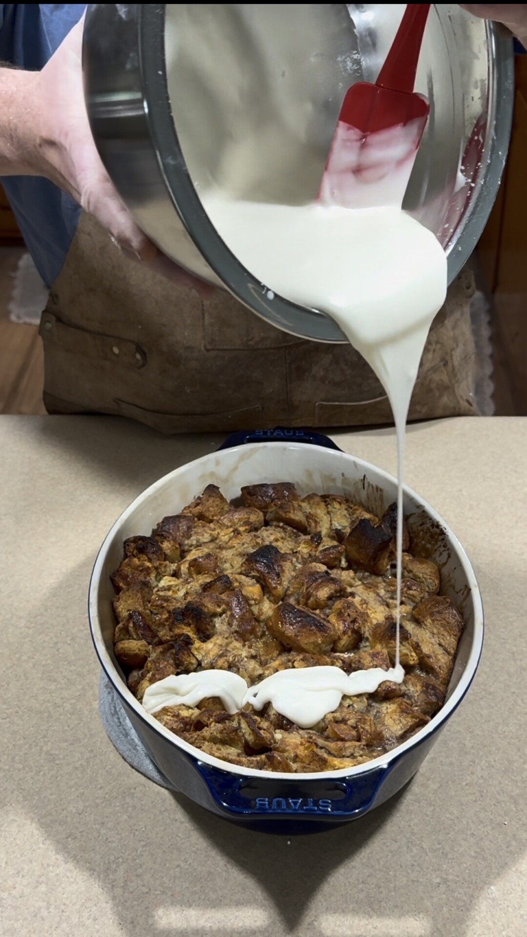 A person pours creamy white sauce from a bowl onto cooked bread pudding in a blue baking dish on a kitchen counter.