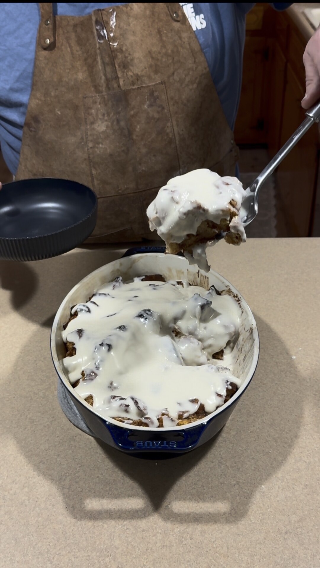 A person serves iced cinnamon rolls from a round baking dish, using a large spoon. The rolls are covered in white icing and placed on a kitchen countertop.