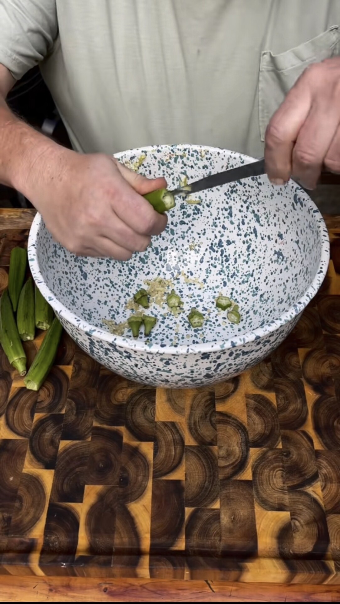 A person slices fresh okra into a speckled white and blue bowl on a wooden cutting board, with whole okra visible beside the bowl.