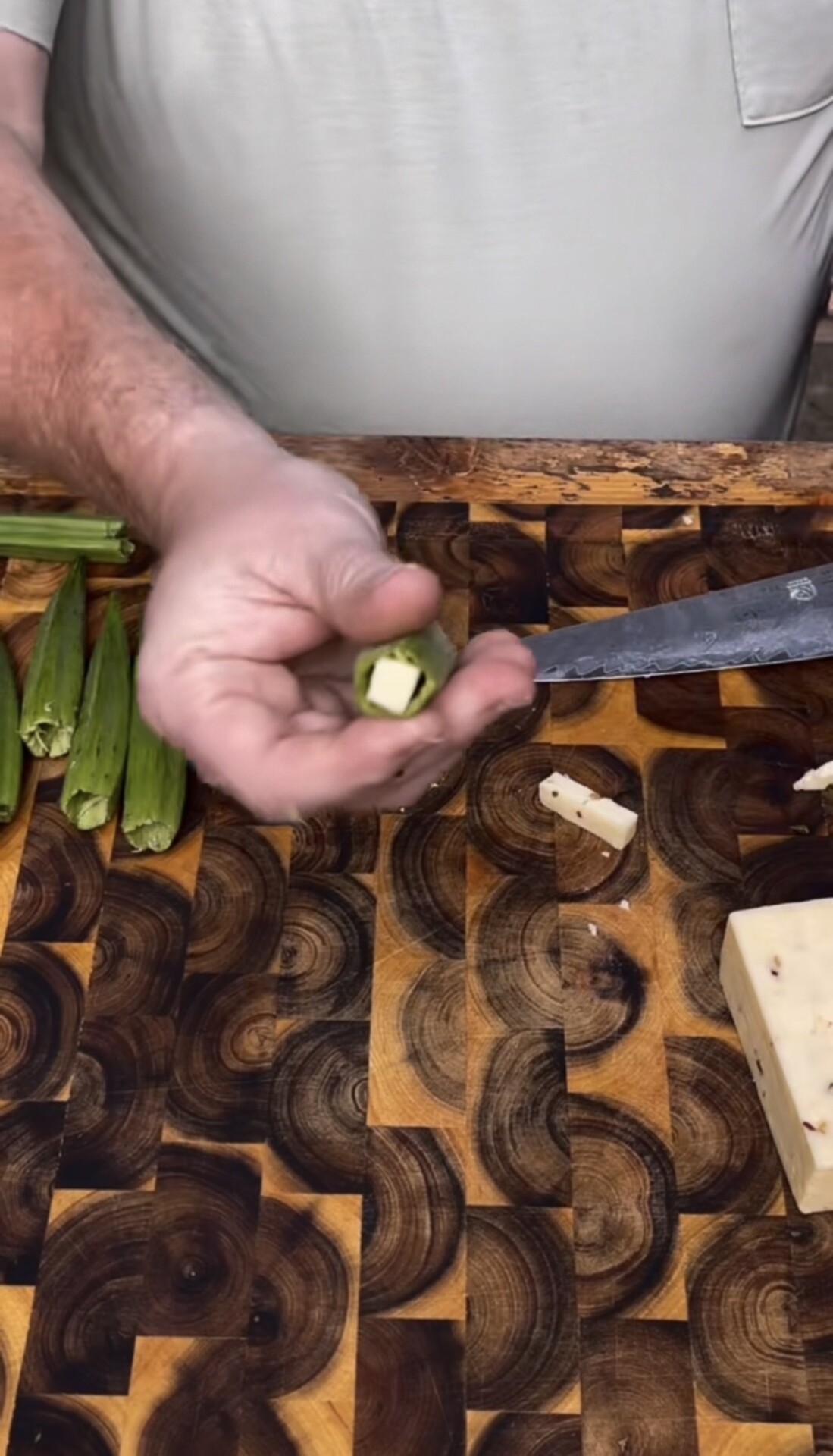 A person holds a sliced piece of okra over a wooden cutting board with a knife, more okra, and a block of cheese.