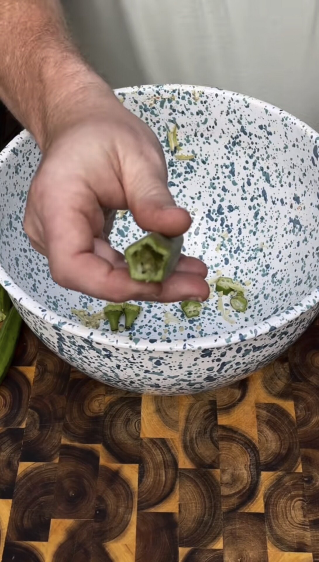 A hand holds a sliced piece of okra above a white speckled bowl with okra pieces inside, on a wooden cutting board.