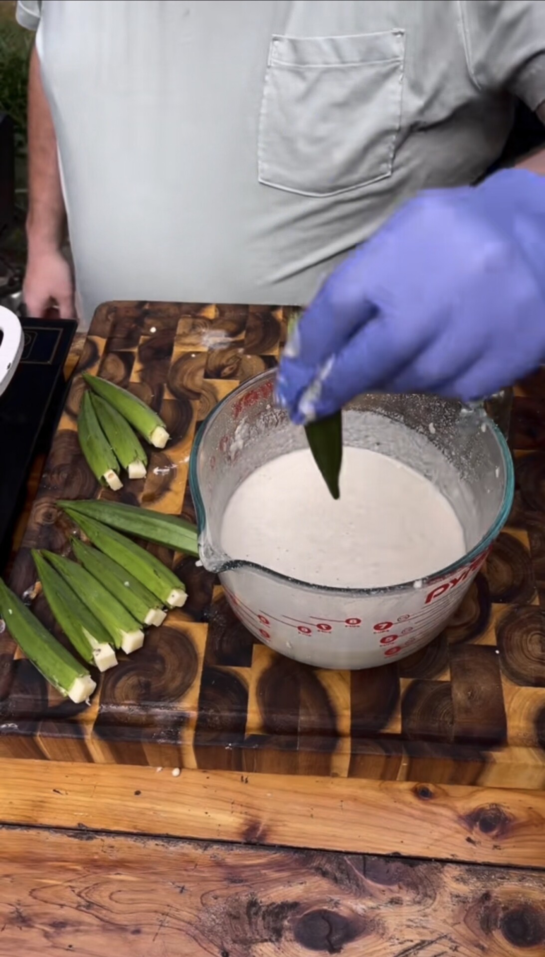 A person wearing a gray shirt and blue glove dips okra into batter on a wooden cutting board, with more okra pieces nearby.