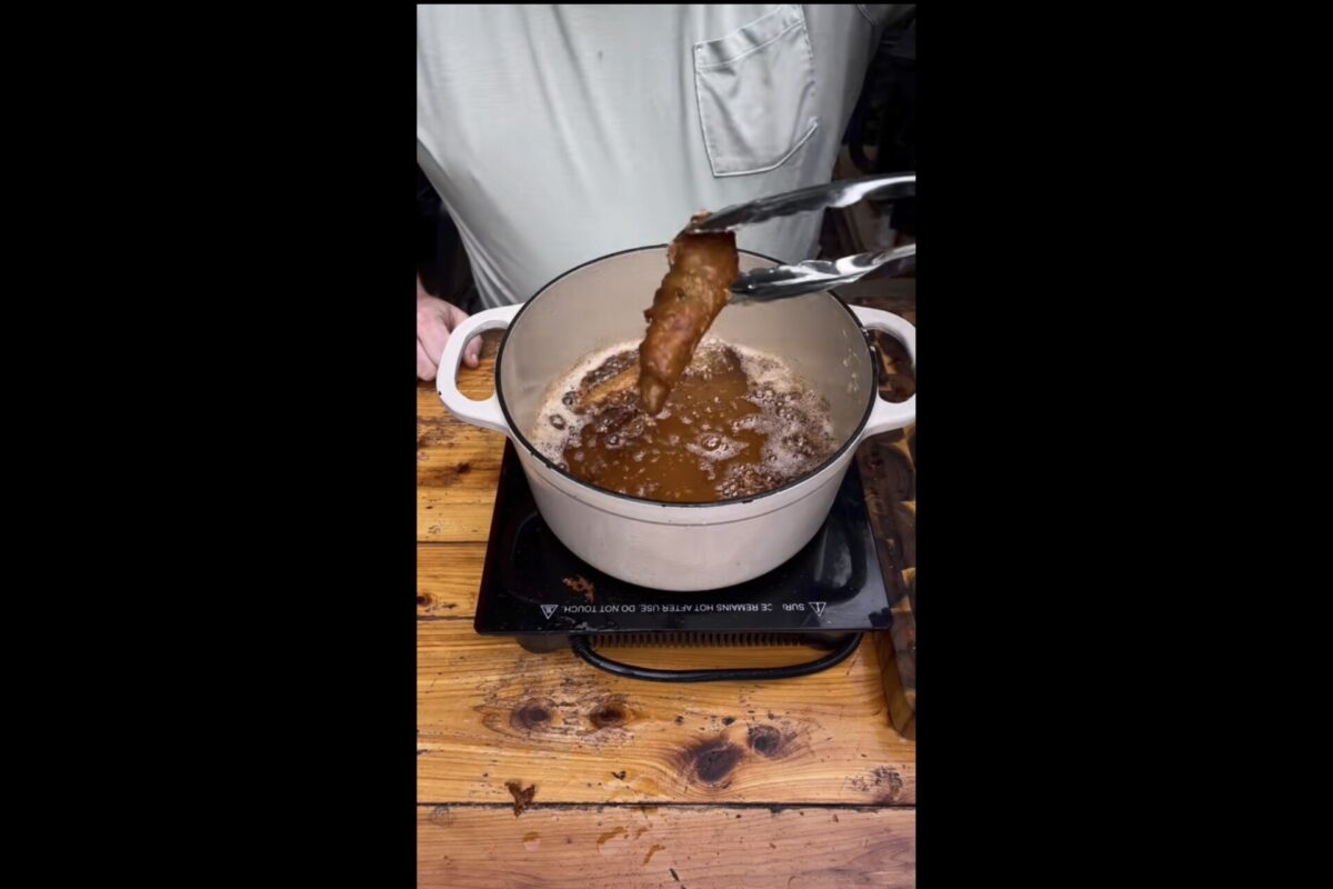 A person uses tongs to lift a piece of fried food from a pot of hot oil on a wooden table, with oil bubbling in the pot.