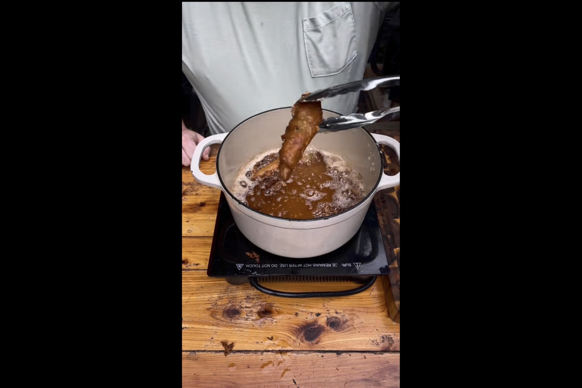 A person uses tongs to lift a piece of fried food from a pot of hot oil on a wooden table, with oil bubbling in the pot.