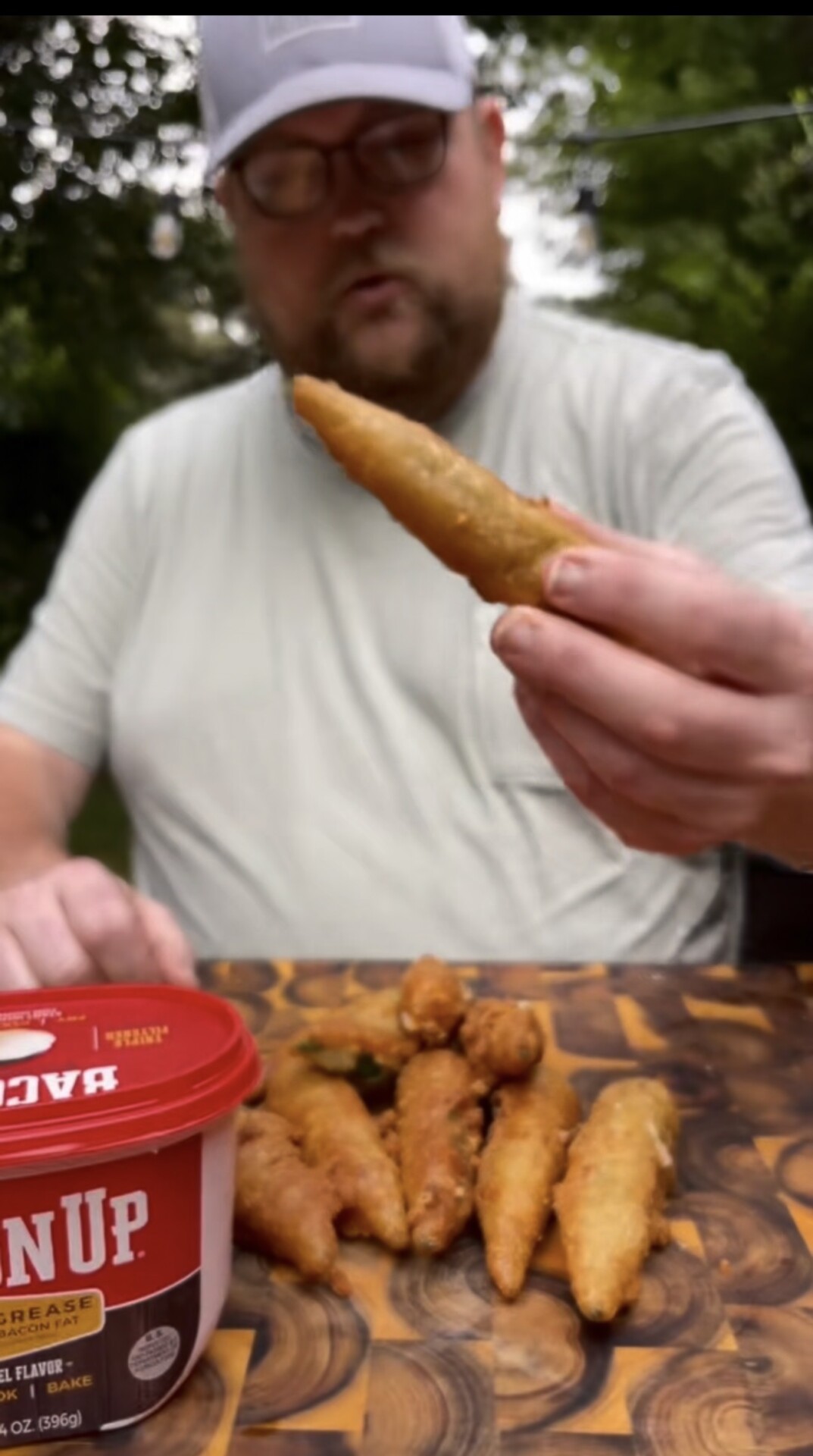 A person holds a piece of fried food above a wooden board with more fried pieces and a container of sauce labeled Ranch outdoors.