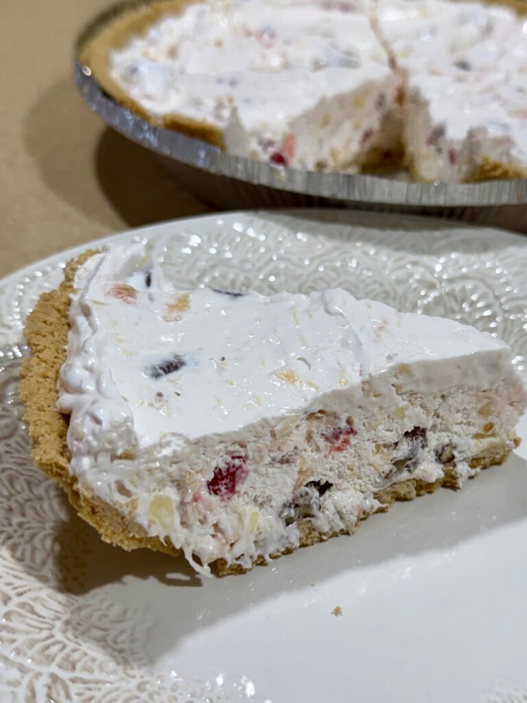 A slice of creamy, fruit-filled pie with a graham cracker crust sits on a white plate. The whole pie, with similar filling, is in the background.