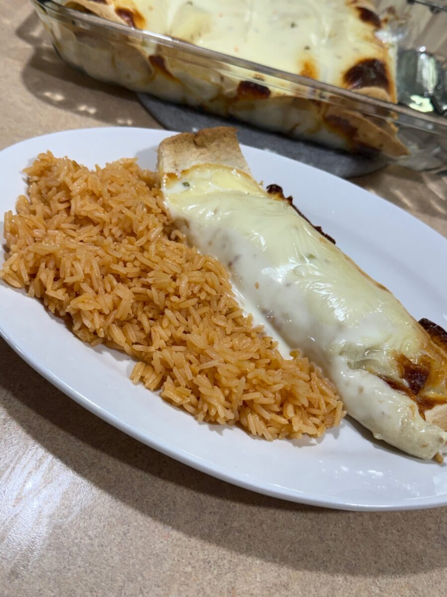 A white plate with orange Mexican rice and a cheesy baked enchilada. In the background, a glass baking dish with more enchiladas topped with melted cheese is visible.