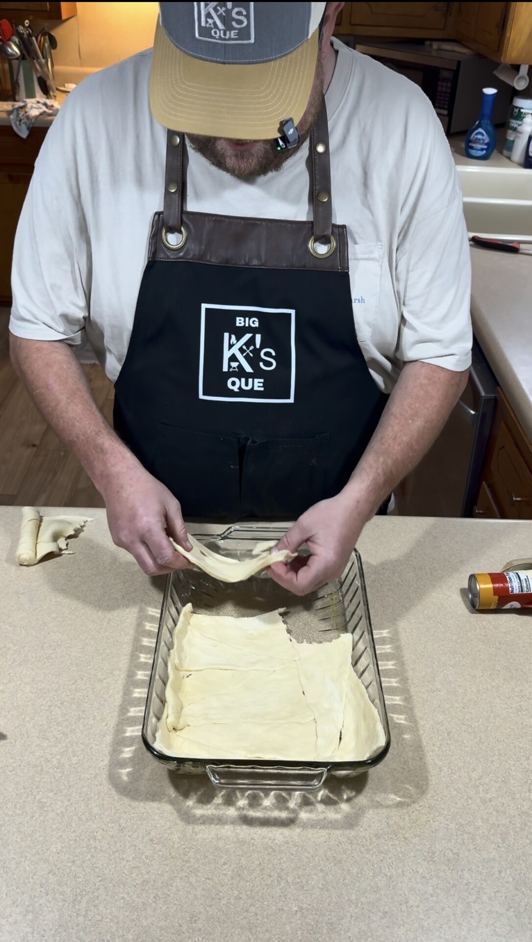 A person wearing a black Big Ks Que apron and tan cap is placing dough sheets into a glass baking dish on a kitchen counter. A can of cooking spray sits nearby.