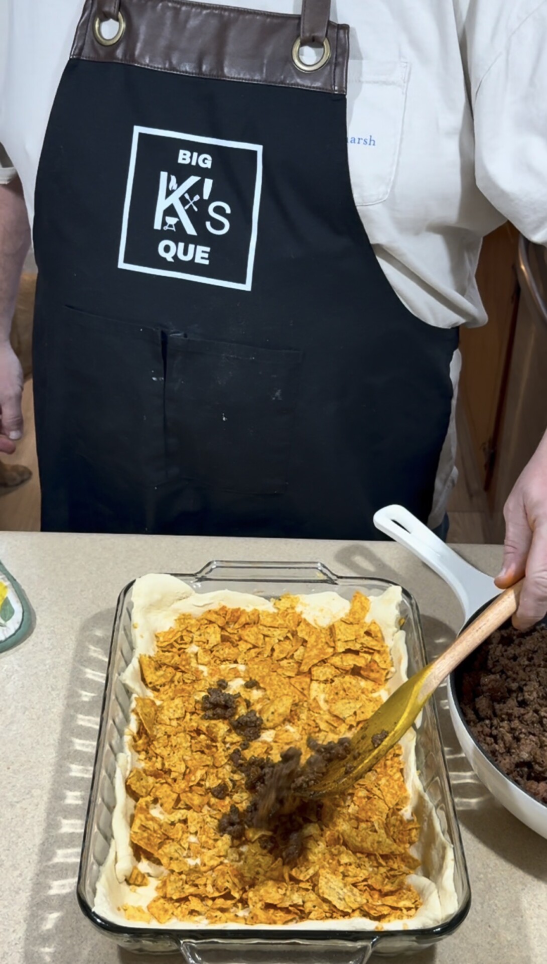 A person wearing a Big Ks Que apron spreads ground beef over a layer of crushed chips in a glass baking dish lined with dough on a kitchen counter.