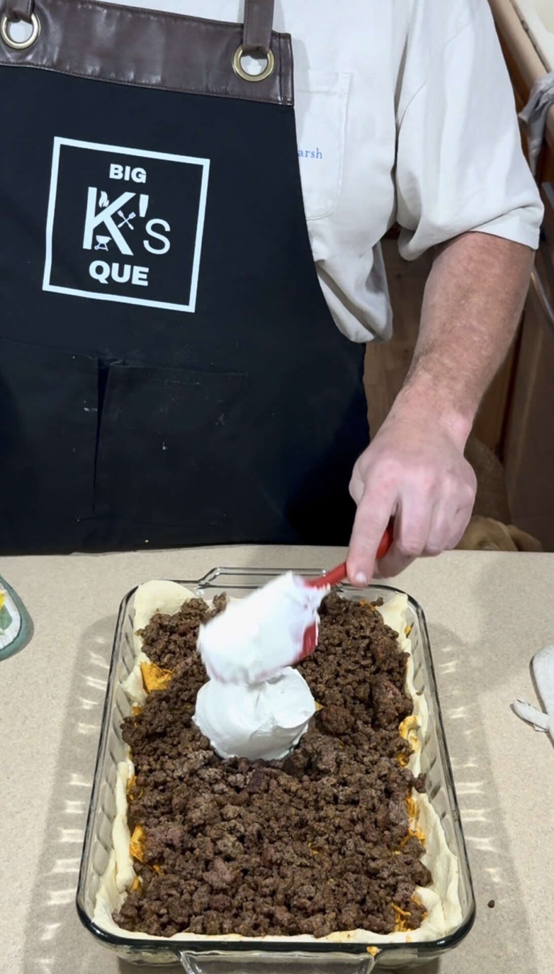 A person in a black apron adds a dollop of sour cream to a casserole dish filled with ground beef and pastry, preparing a layered dish on a kitchen counter.
