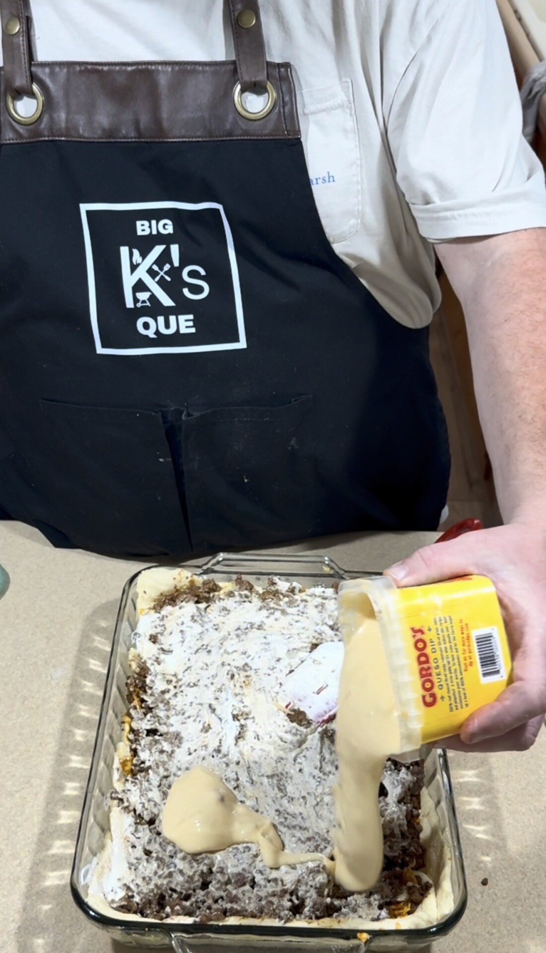 A person wearing a black apron labeled BIG KS QUE spreads cheese sauce from a jar labeled Gordos over a layered casserole in a glass dish.