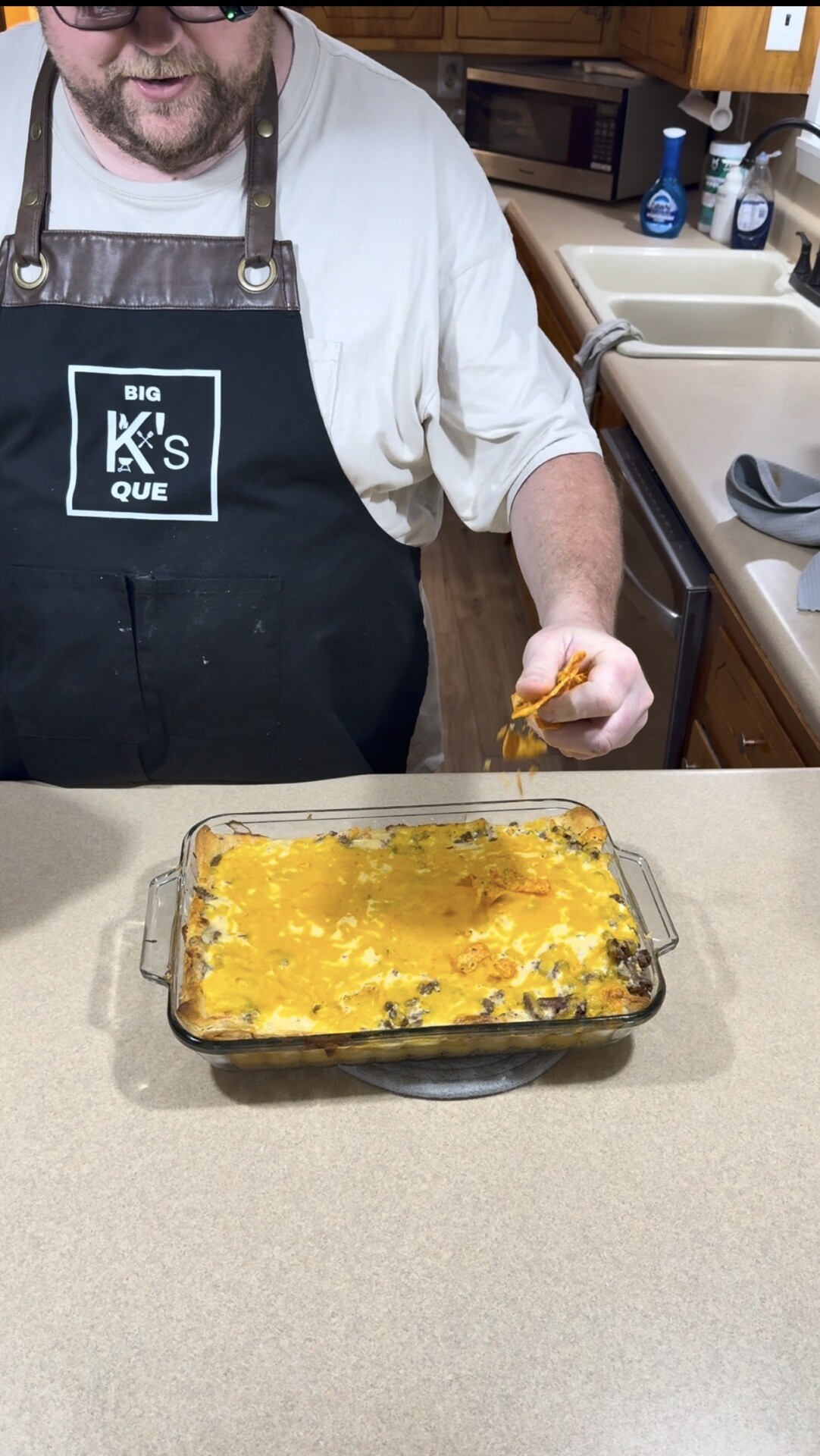 A person in an apron stands by a kitchen counter, holding a cheesy serving from a glass dish filled with baked casserole. The kitchen has wooden cabinets and a sink visible in the background.
