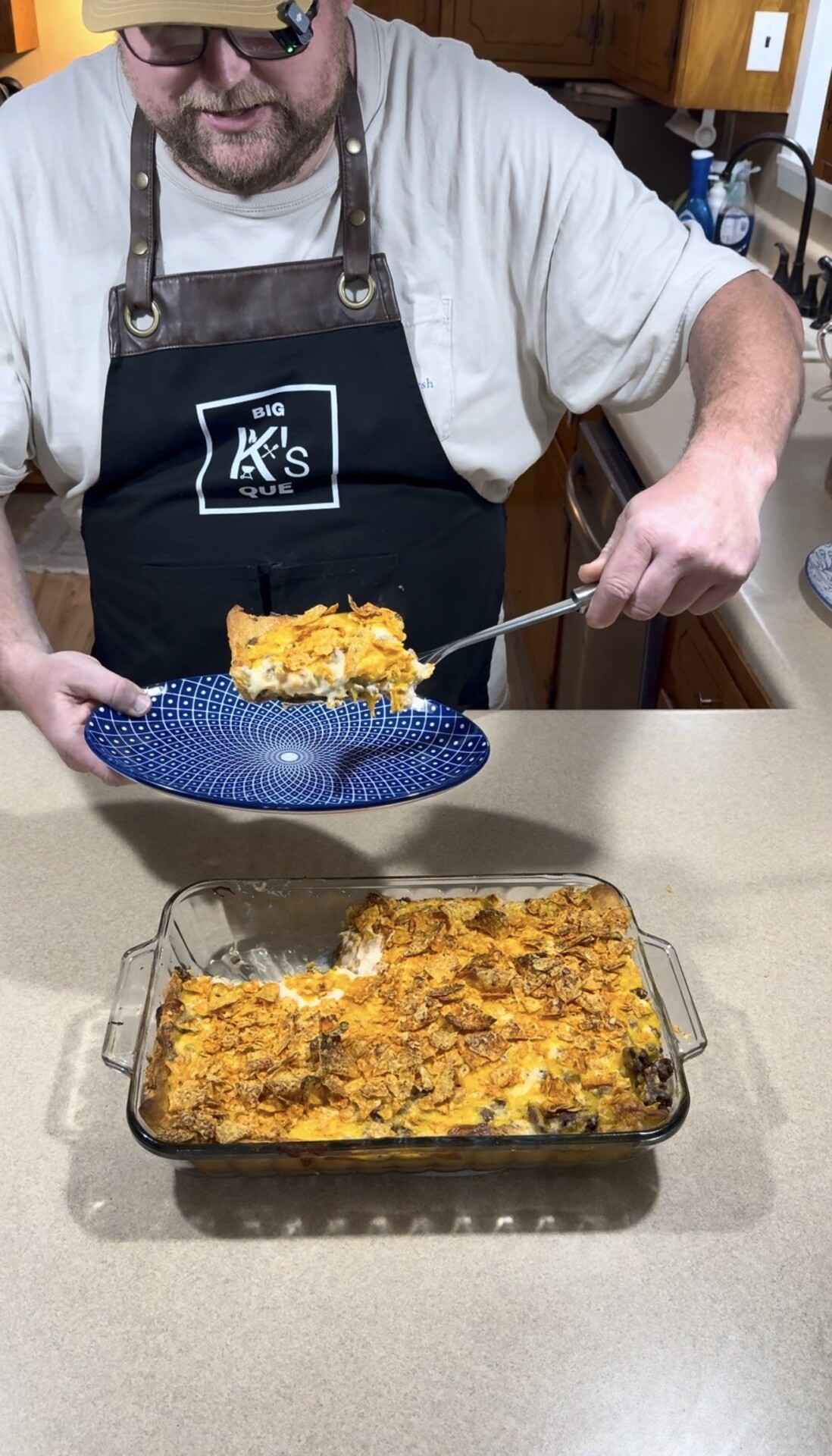A person in an apron serves a cheesy casserole with a crunchy topping onto a blue plate from a glass baking dish on a beige kitchen counter.