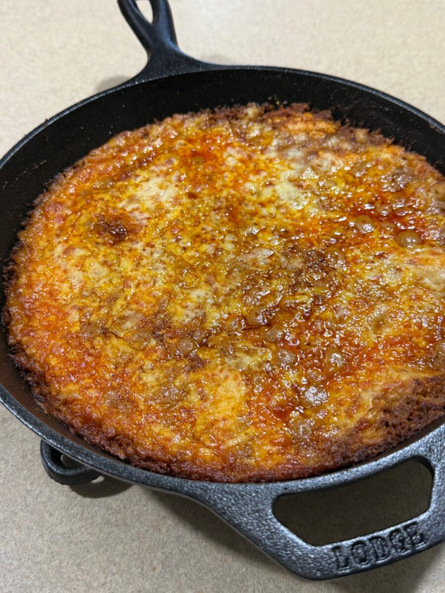 A cheesy, golden-brown deep dish pizza in a black cast iron skillet sits on a beige countertop.