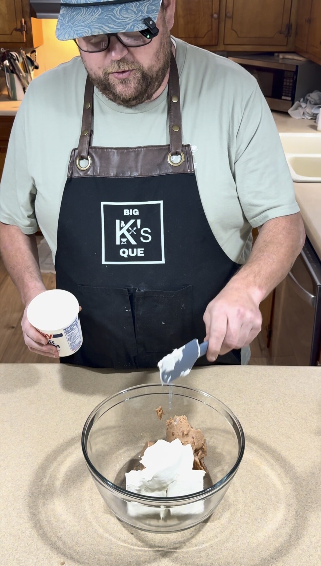 A person in a black apron adds a white ingredient from a container into a glass bowl with brown and white mixtures on a kitchen counter.