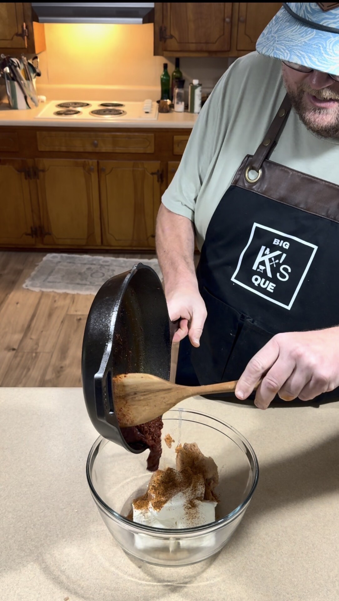 A man in an apron pours cooked meat from a skillet into a glass bowl containing cream cheese and seasoning on a kitchen counter.