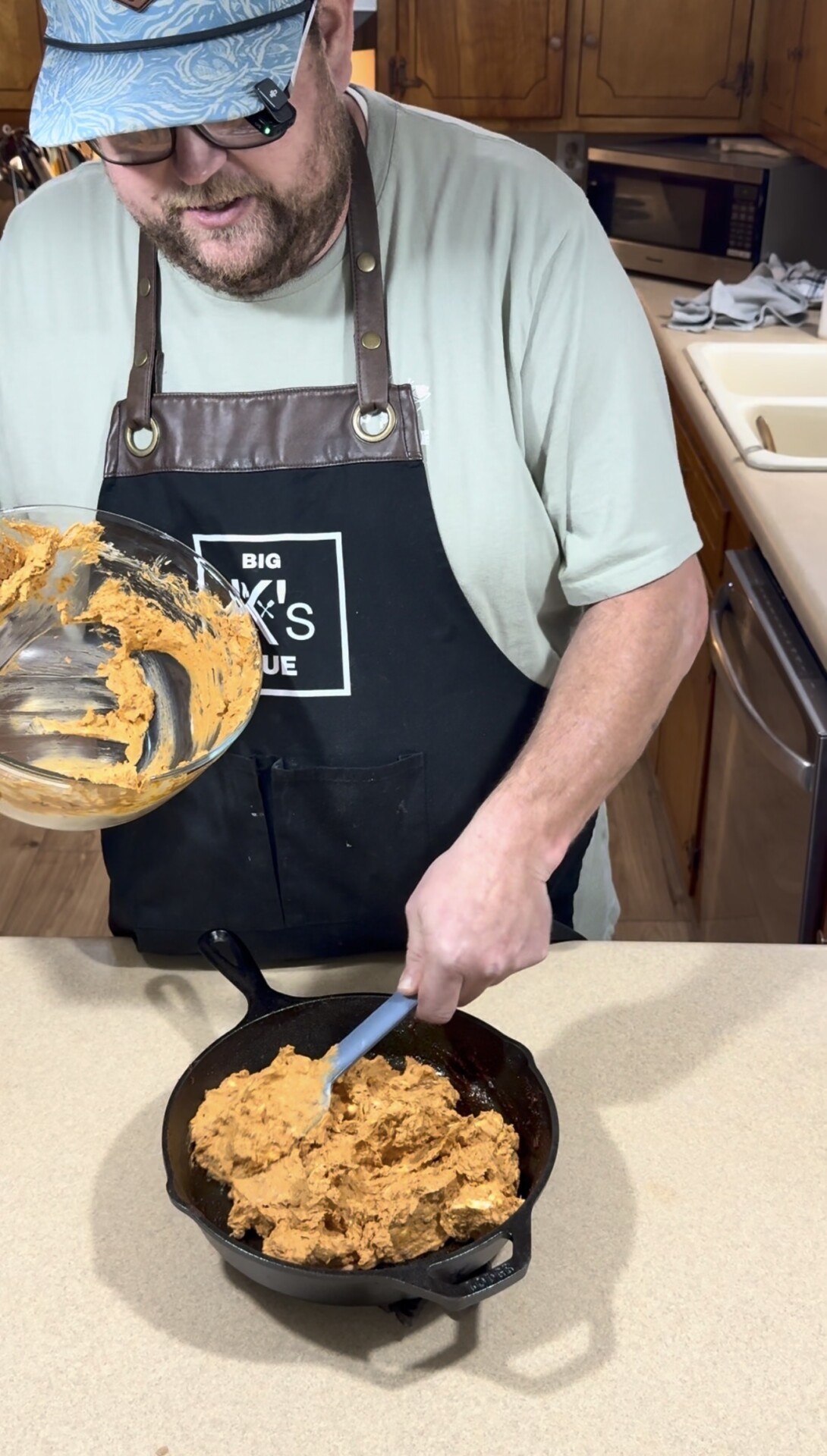 A person wearing an apron spreads a thick orange batter into a black cast iron skillet on a kitchen counter, holding a mixing bowl and spatula.