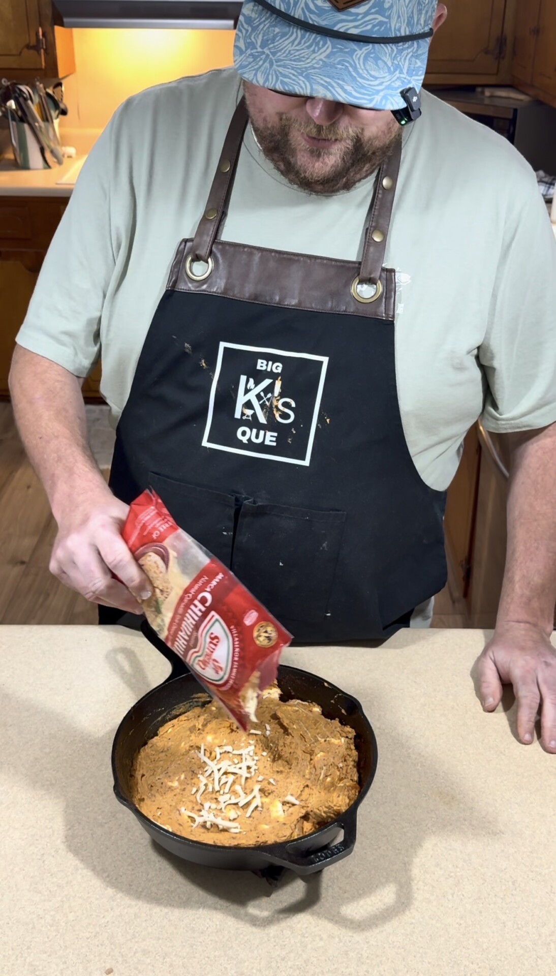 A person in an apron pours shredded cheese from a bag into a cast iron skillet containing a cheesy dip, standing in a kitchen.