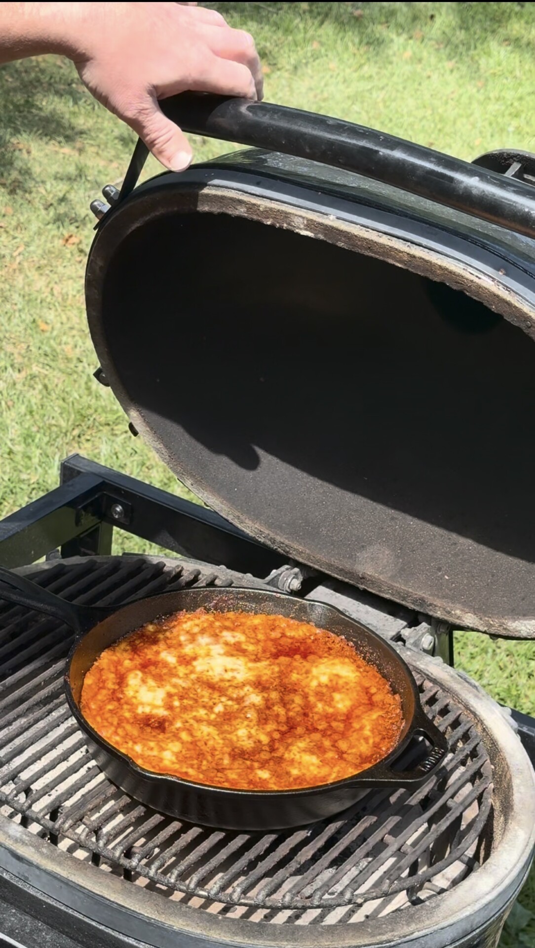 A person lifts the lid of a black grill, revealing a cast iron skillet filled with bubbling, red-orange sauce and melted cheese, outdoors on a sunny day with green grass in the background.