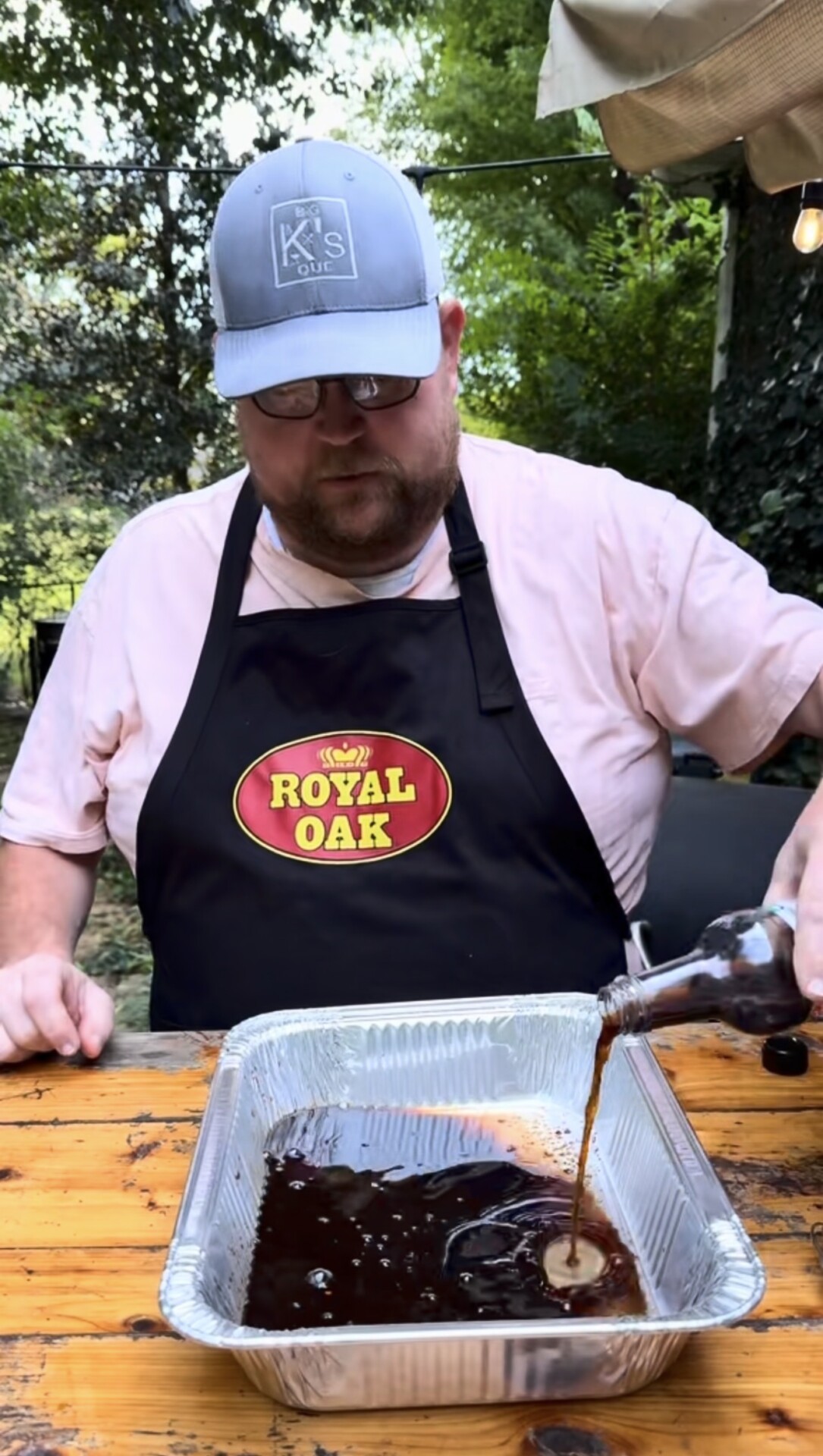 A man wearing a Royal Oak apron and a gray cap pours a brown liquid from a bottle into a foil tray on a wooden table outdoors, surrounded by greenery.