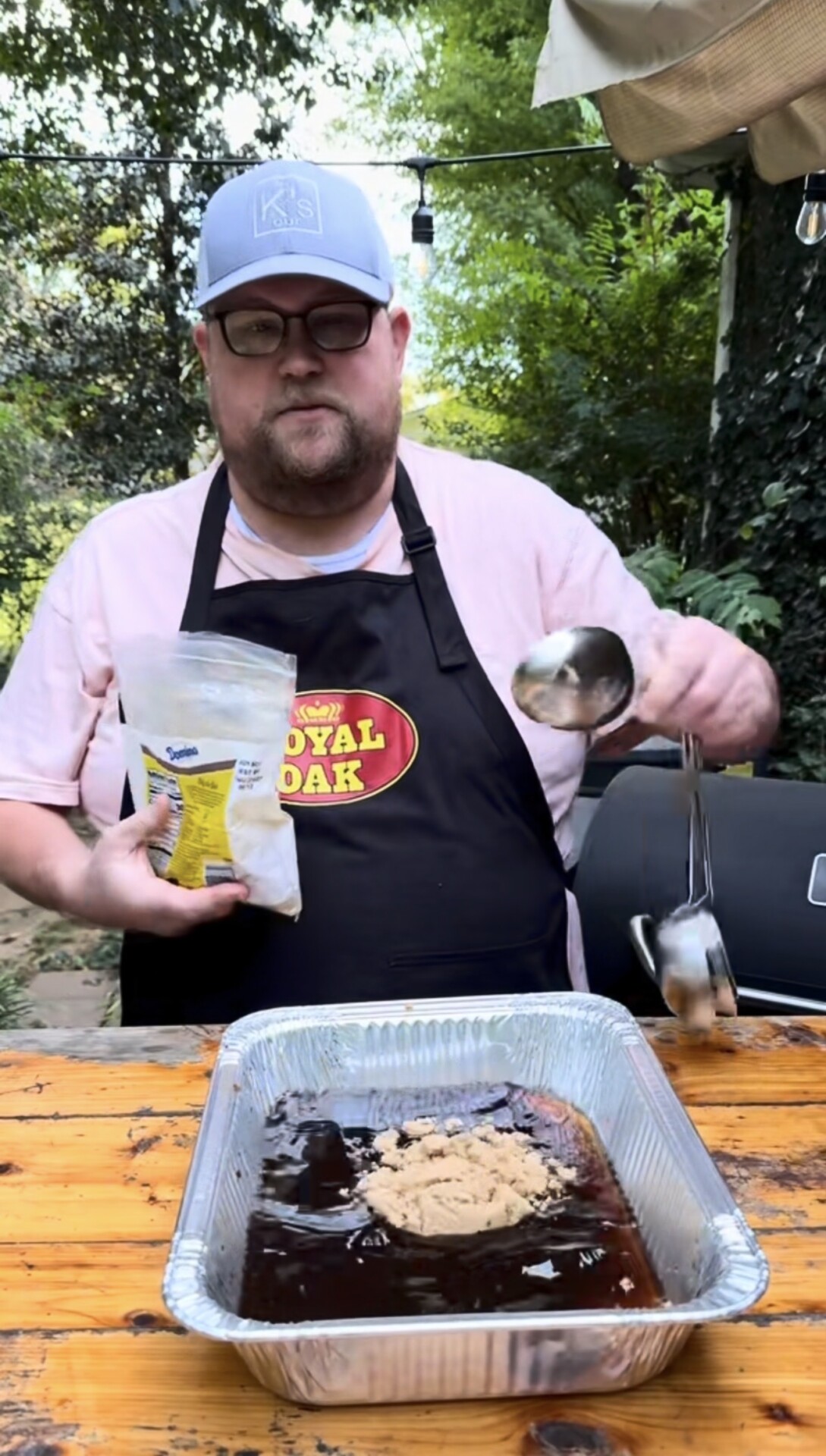 A person in a black apron adds brown sugar from a bag into a foil tray with sauce on a wooden table outdoors, near a grill. Trees and greenery are in the background.
