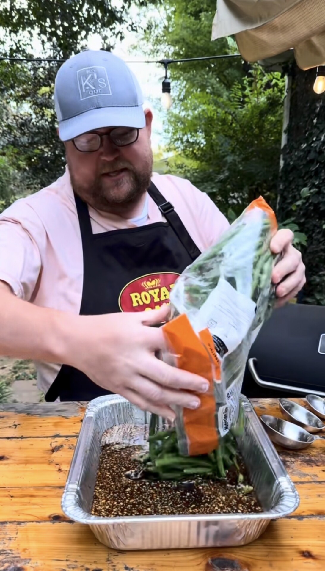 A man wearing a cap and apron pours a bag of green beans into a foil tray with marinade on a wooden outdoor table, surrounded by greenery.