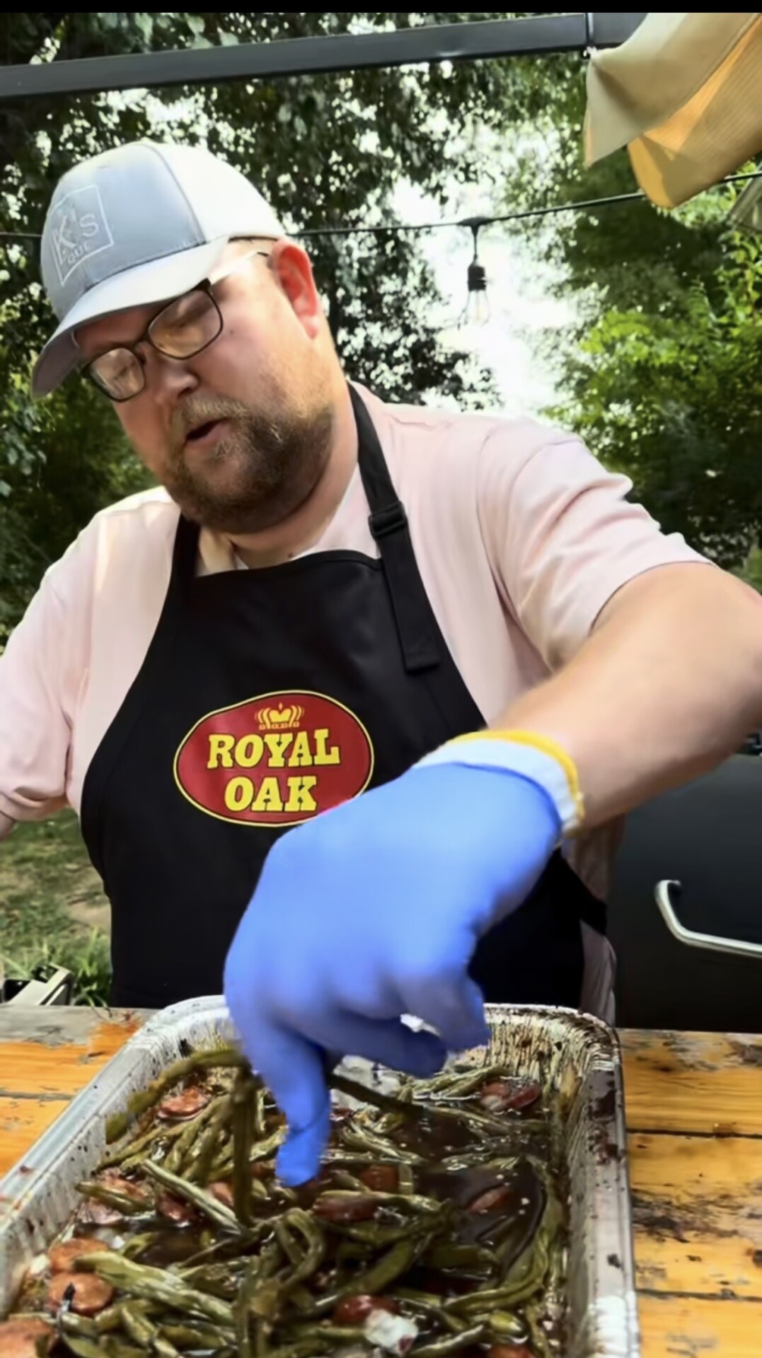 A man wearing a Royal Oak apron and blue gloves prepares green beans and sausage in a tray outdoors, with trees and string lights in the background.
