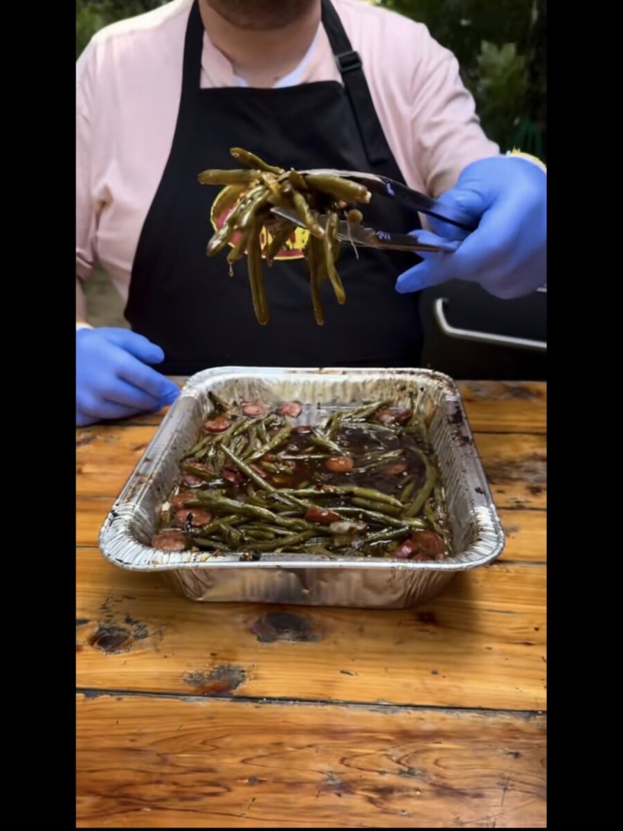 A person in blue gloves and a black apron uses tongs to lift cooked green beans with bacon from a foil pan on a wooden table outdoors.