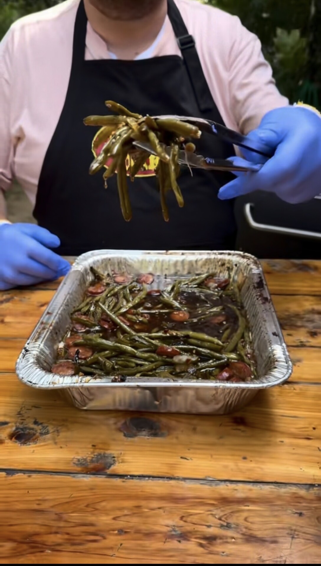 A person wearing blue gloves and a black apron serves cooked green beans from a foil tray using tongs. The tray sits on a wooden table outdoors.