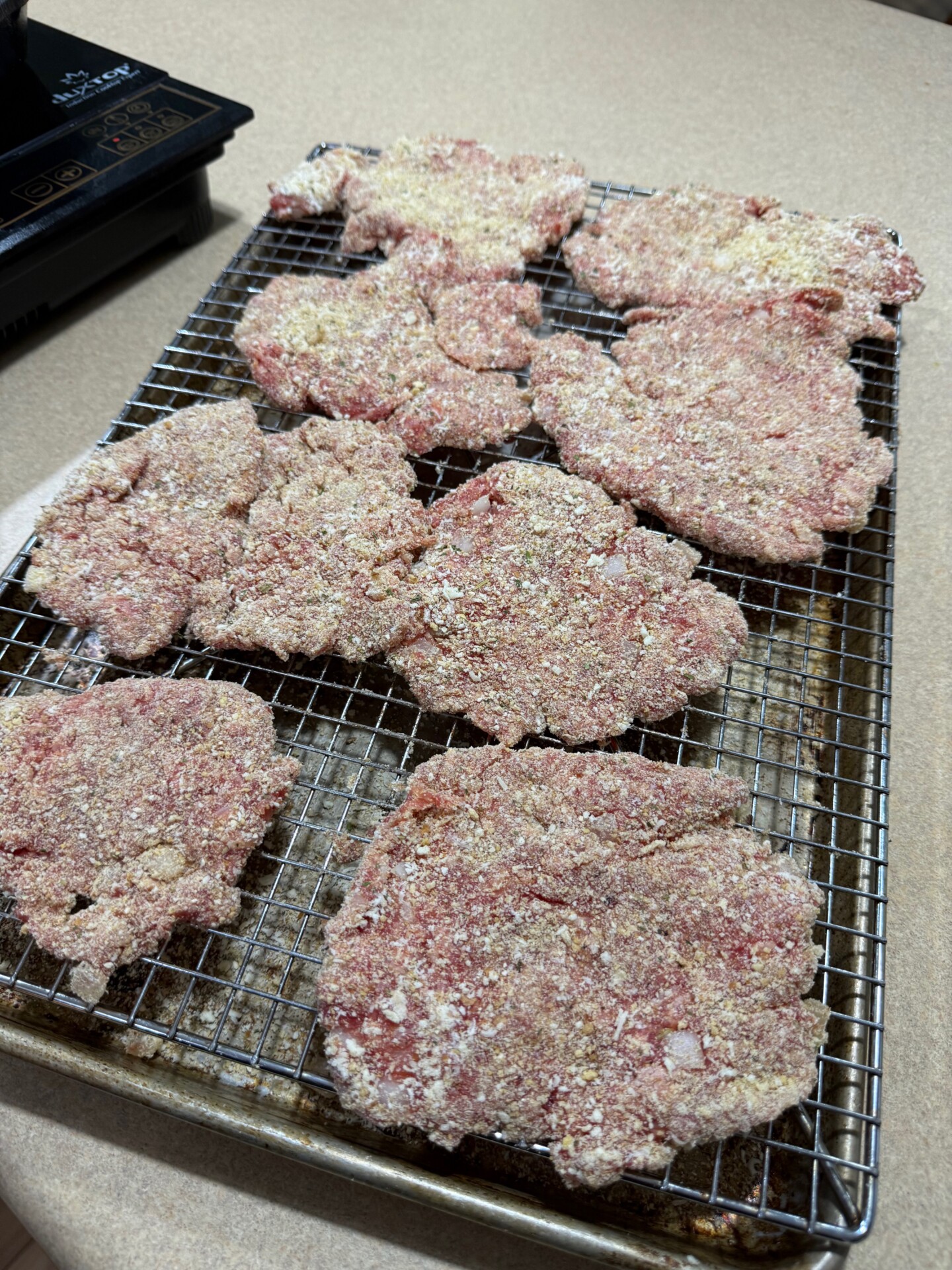 Several uncooked, breaded meat patties are arranged on a wire rack set over a baking sheet, resting on a beige countertop.