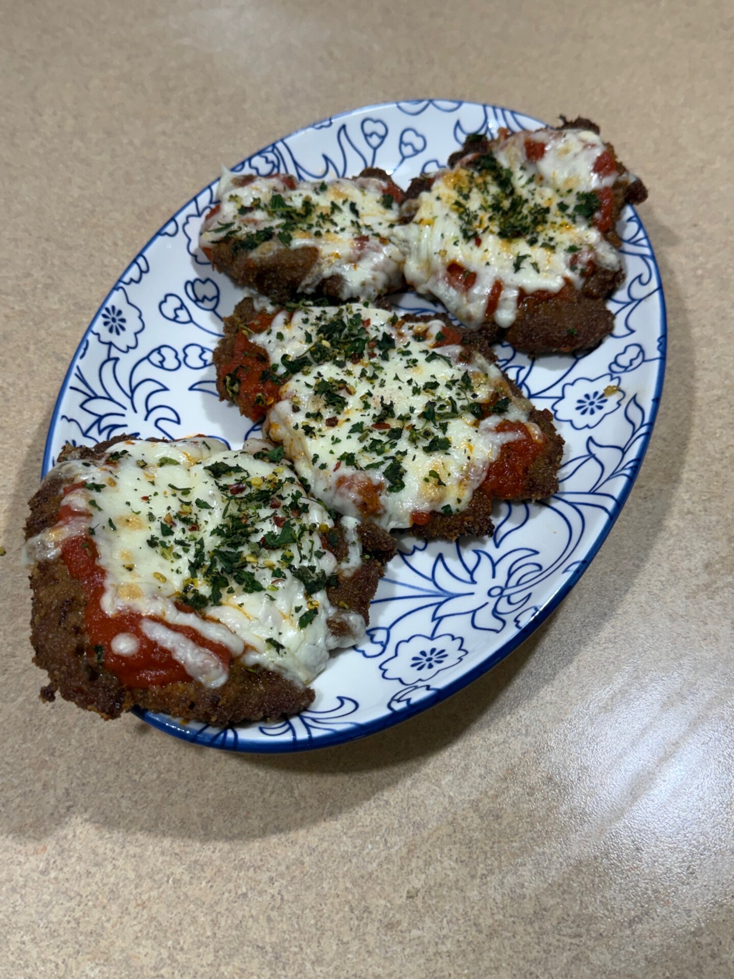 Four breaded and baked chicken cutlets topped with marinara sauce, melted cheese, and chopped herbs are arranged on an oval, blue-and-white floral plate, set on a tan countertop.
